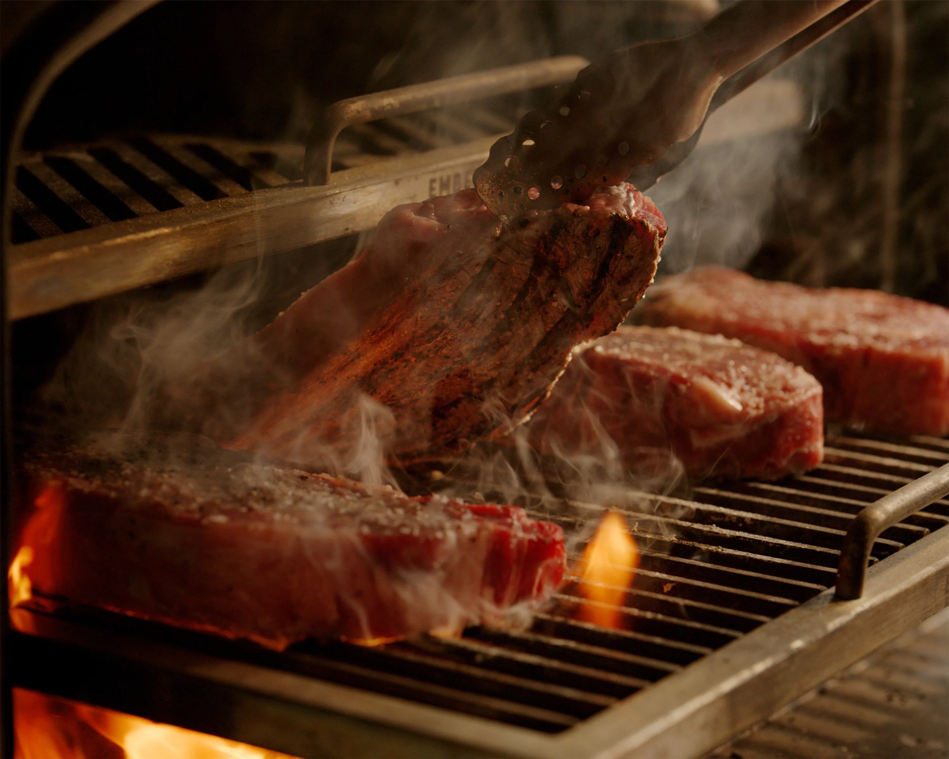 Four thick raw steaks cooking on a metal grill with flames and smoke, one being flipped with tongs.