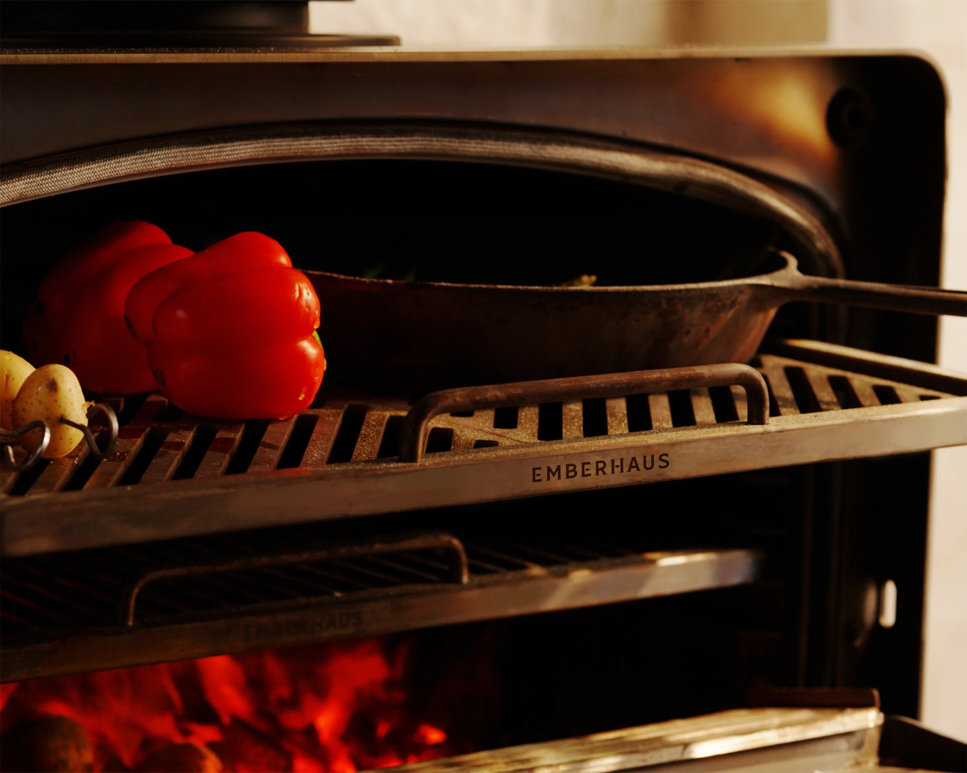 Cast iron skillet and red bell peppers on a metal grill rack labeled "EMBERHAUS" inside a wood-fired oven.