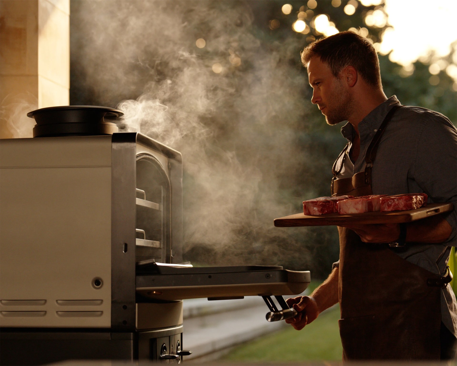 Man in a brown apron holding a wooden board with raw steaks, opening a steaming outdoor oven.