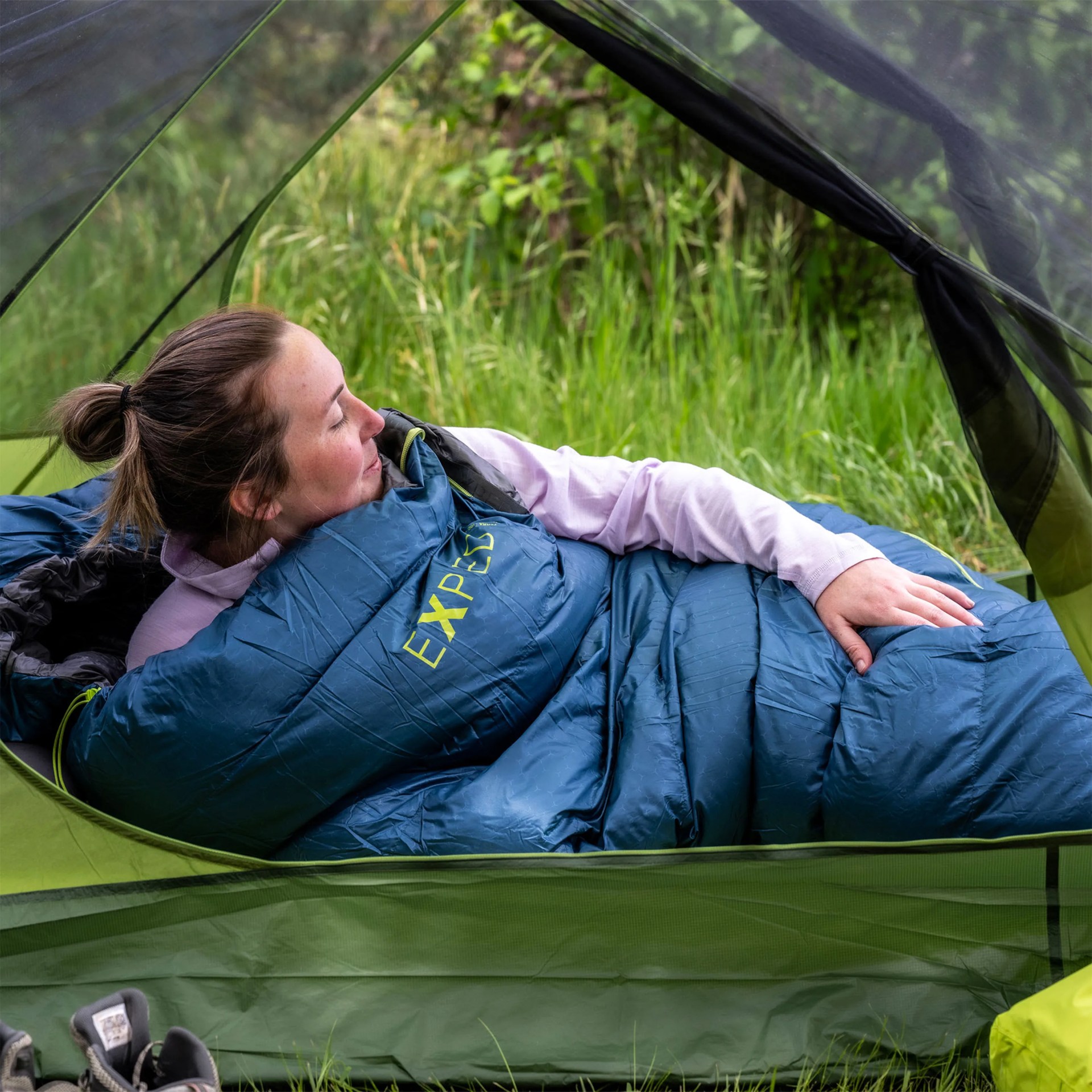 Person lying inside a green tent wrapped in a blue EXPED sleeping bag on grass.