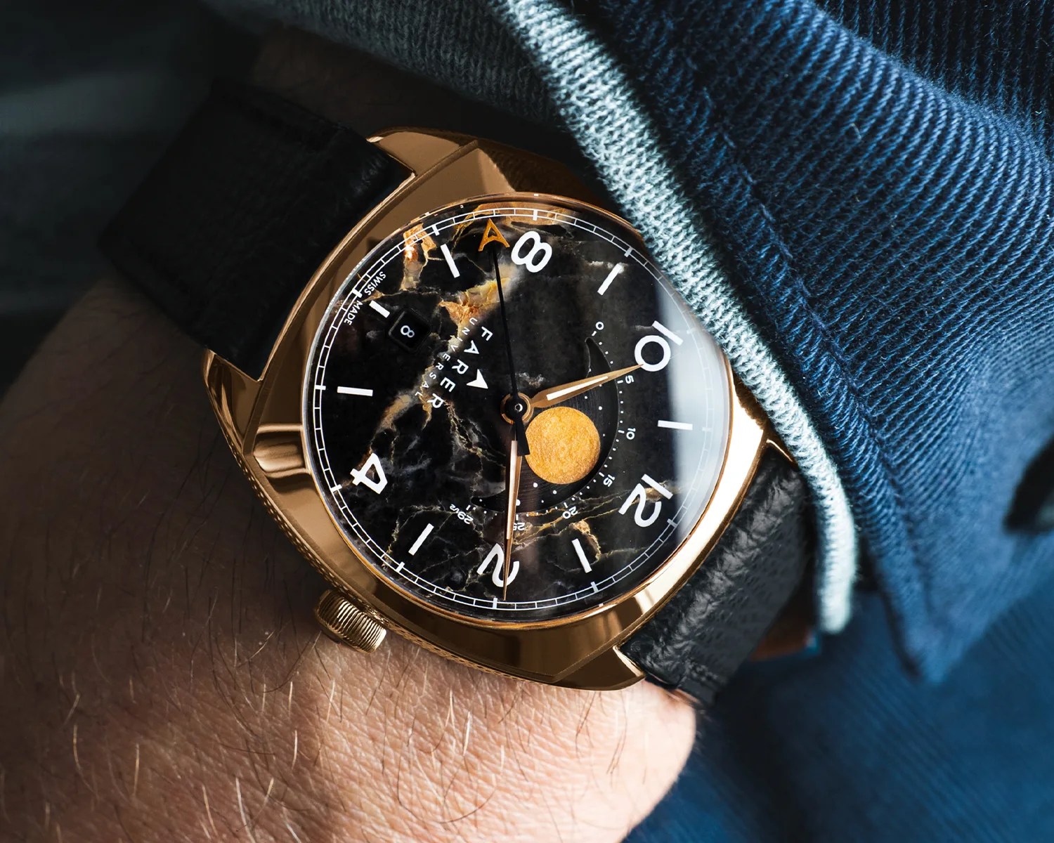 Gold-toned wristwatch with black marbled dial, white numerals, and black leather strap worn under a blue sleeve.