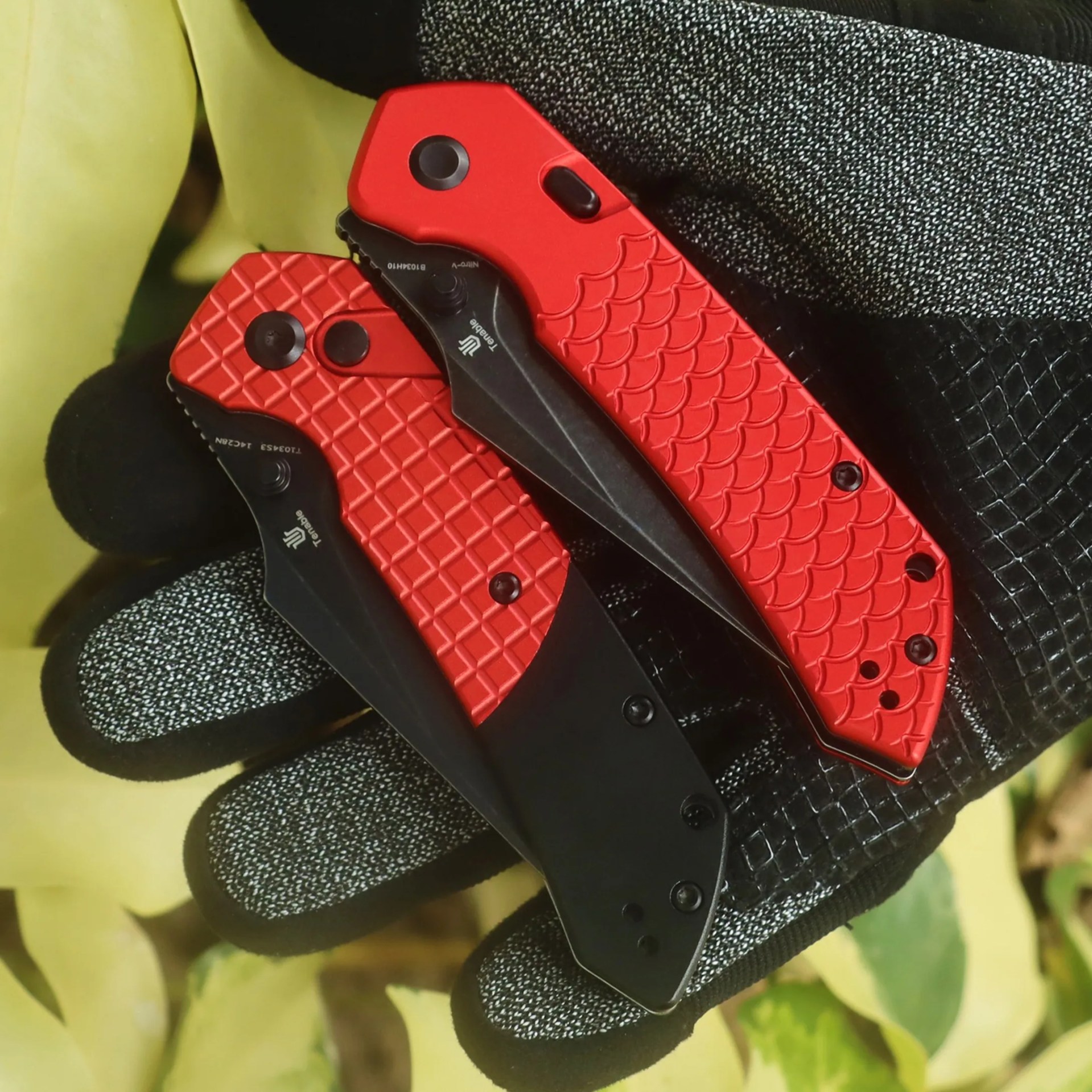 Two red and black folding knives with textured handles held in a gloved hand against a leafy background.
