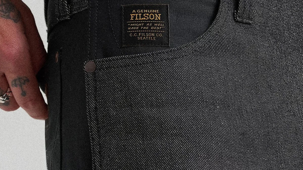 Close-up of black denim pants pocket with a black and gold Filson brand label and a tattooed hand nearby.