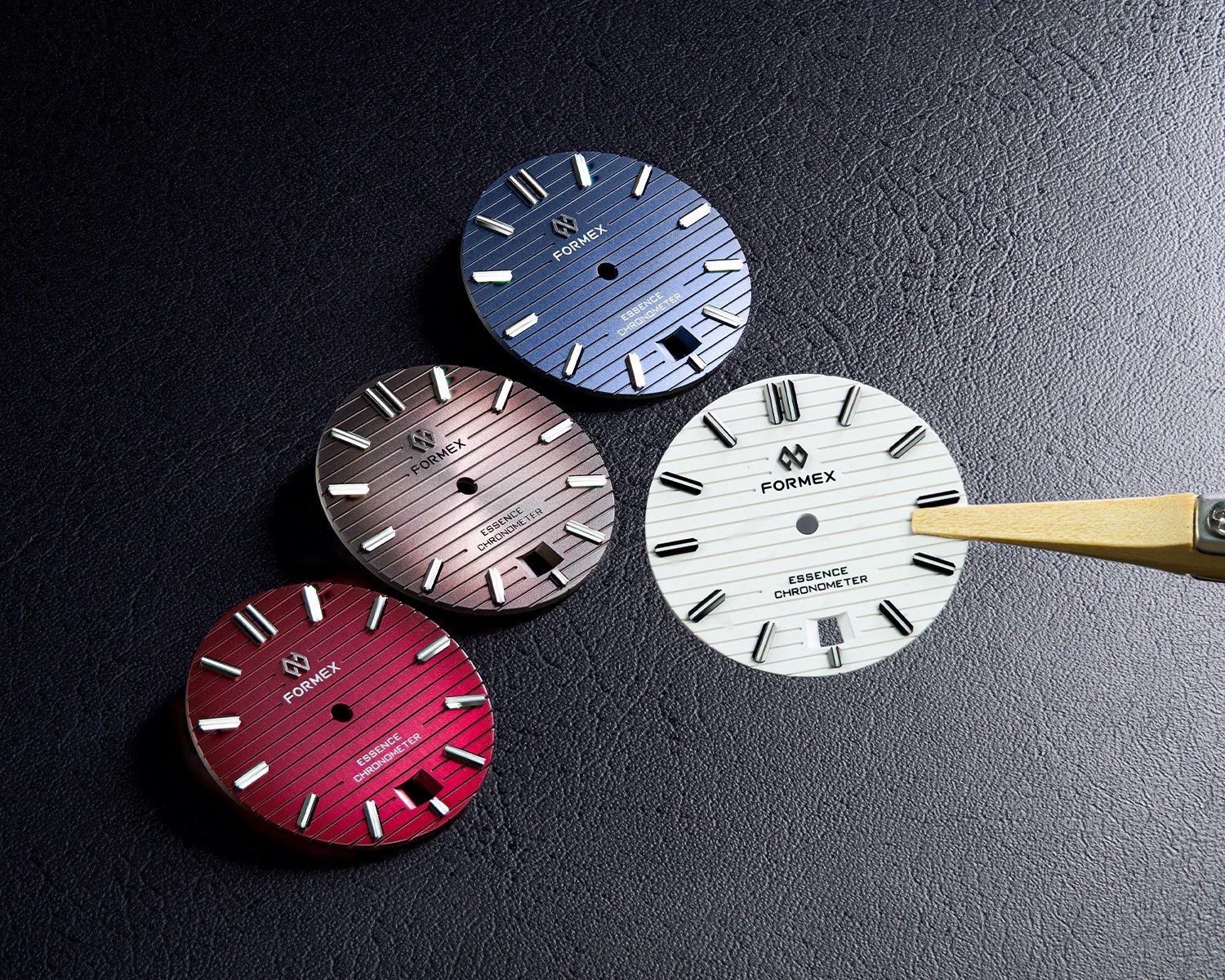 Four round Formex Essence Chronometer watch dials in blue, brown, red, and white on a black textured surface with a wooden tool pointing at the white dial.