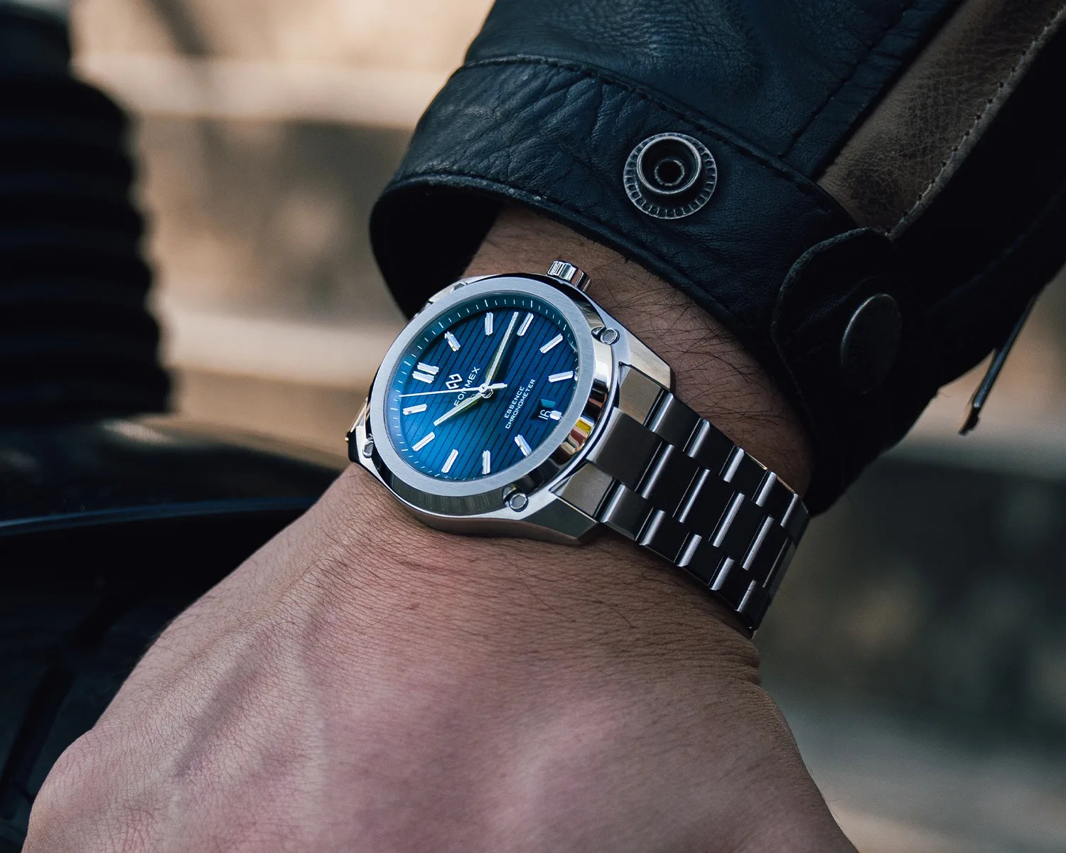 Silver metal wristwatch with a blue textured dial and white hour markers worn on a wrist with a black leather jacket.