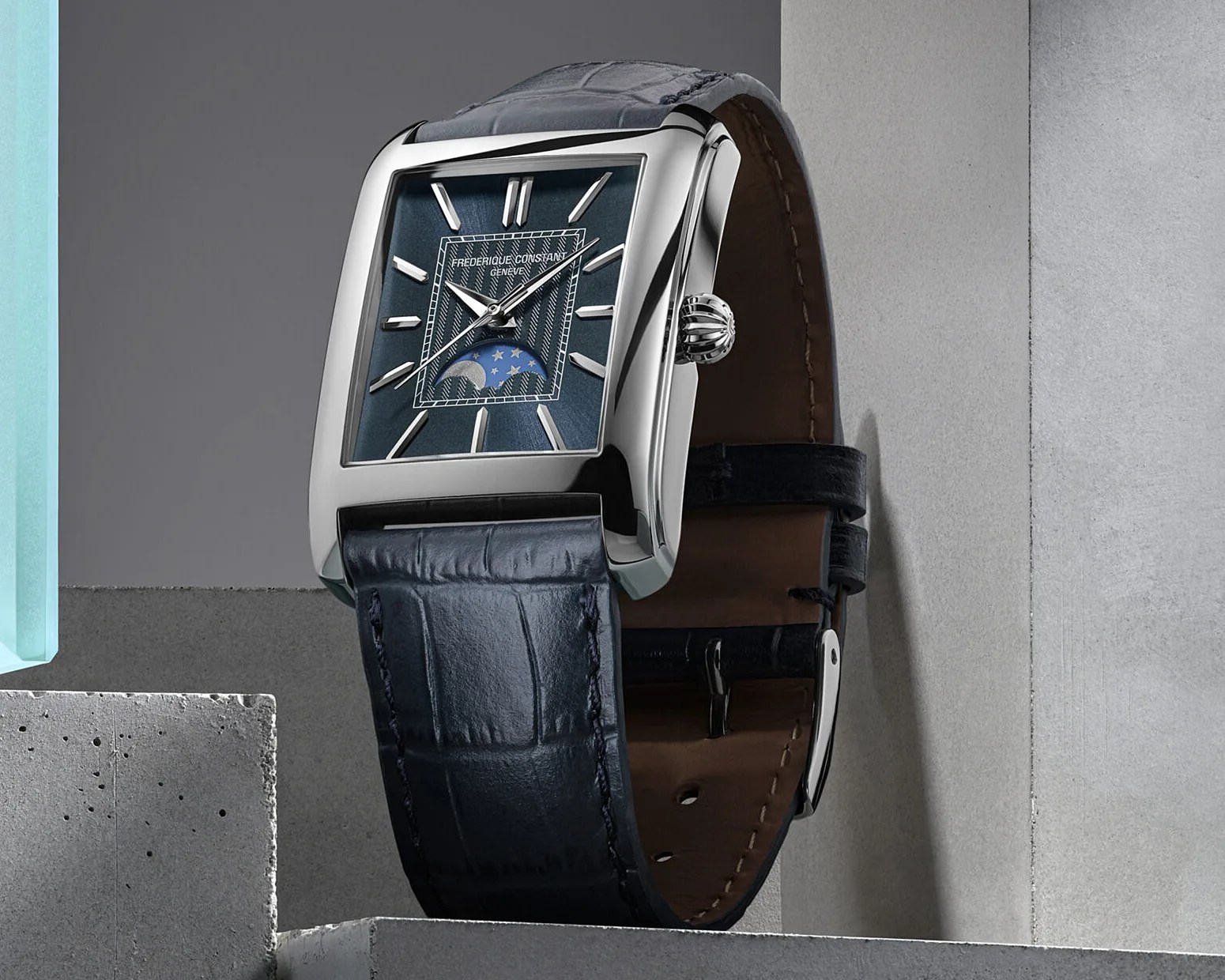 Rectangular silver watch with blue dial, moon phase indicator, and dark blue leather strap on concrete blocks.