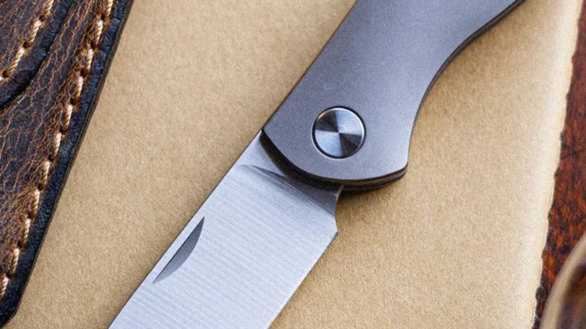 Close-up of a folding knife with a silver blade and gray handle on a beige surface next to brown leather.