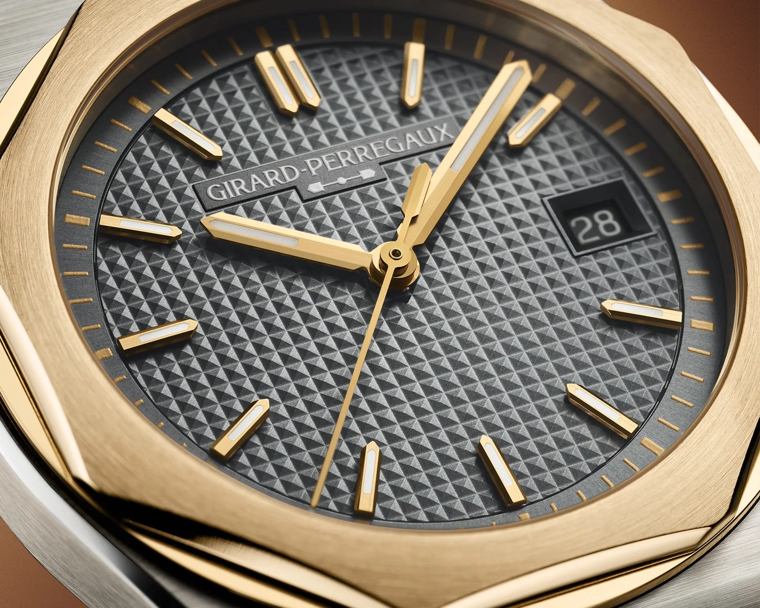 Close-up of a Girard-Perregaux watch face with a textured gray dial, gold hour markers, hands, and a date window showing 28.