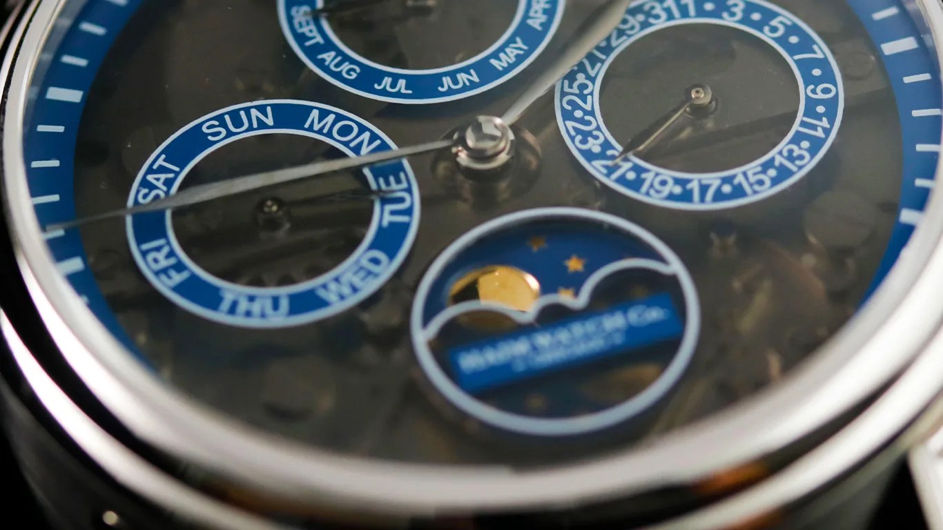 Close-up of a watch face showing day, date, month, and moon phase subdials with blue and white markings.