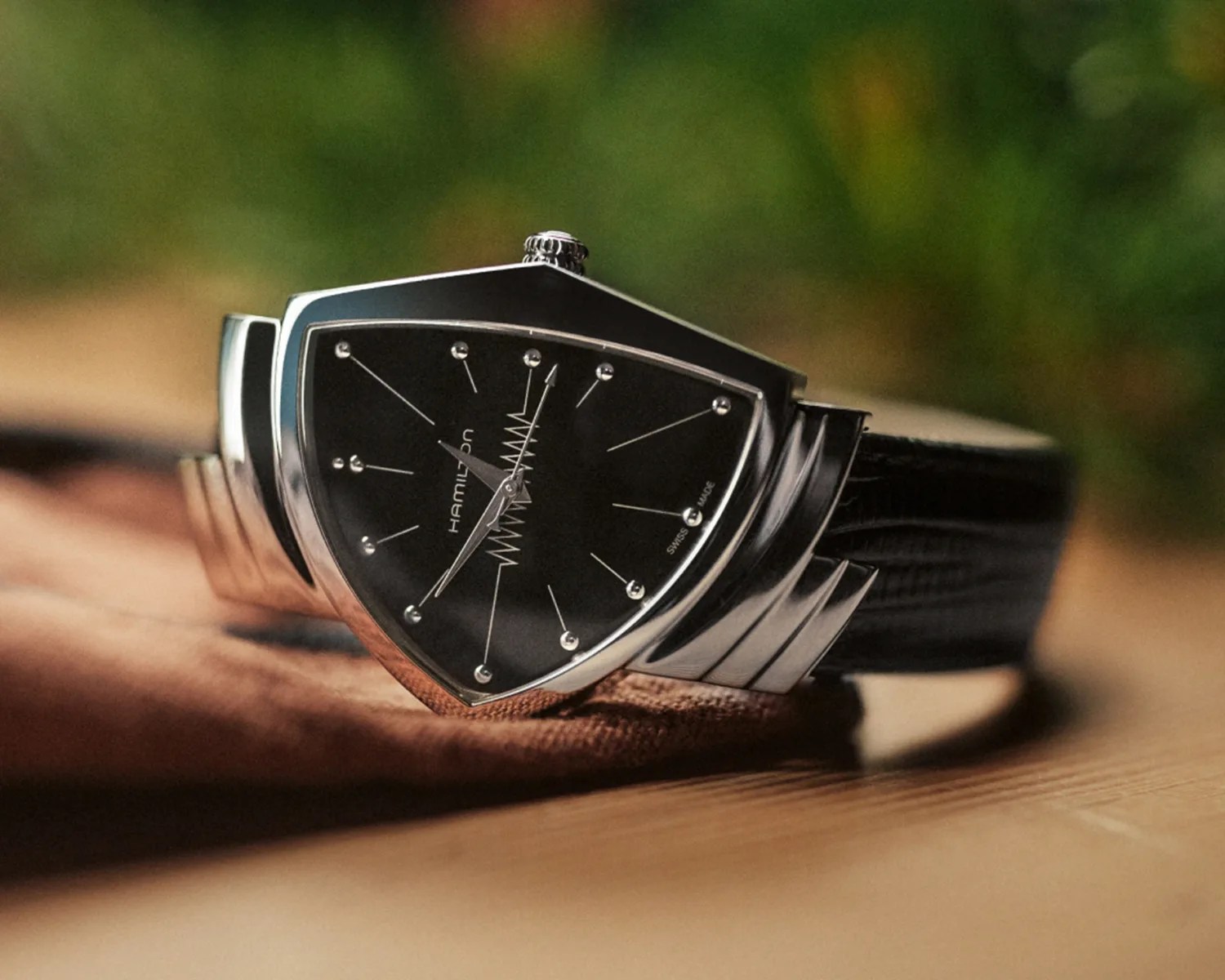 Triangular stainless steel Hamilton wristwatch with black dial and black leather strap on a wooden surface.