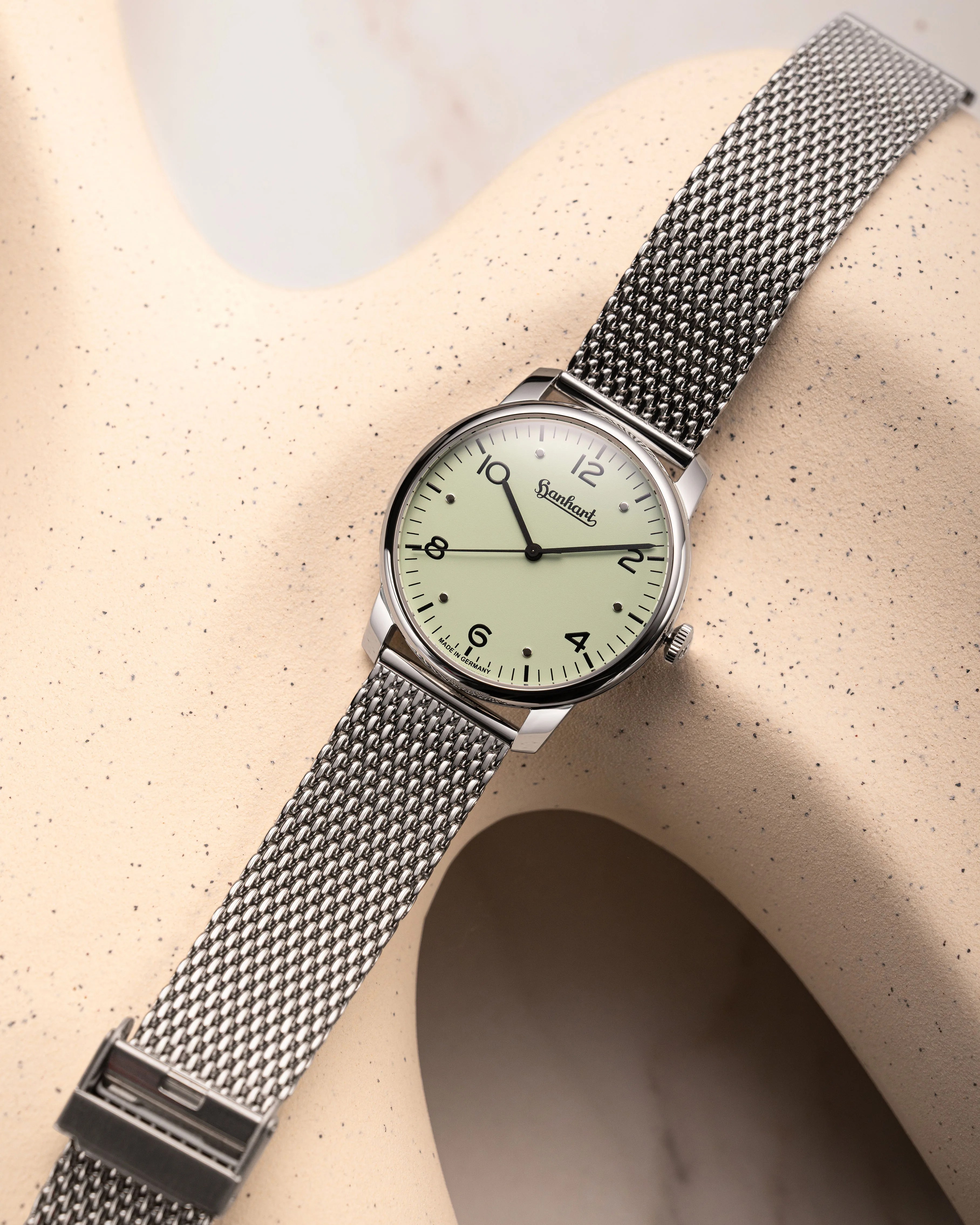 Silver mesh band wristwatch with pale green dial and black numerals on a beige speckled abstract surface.