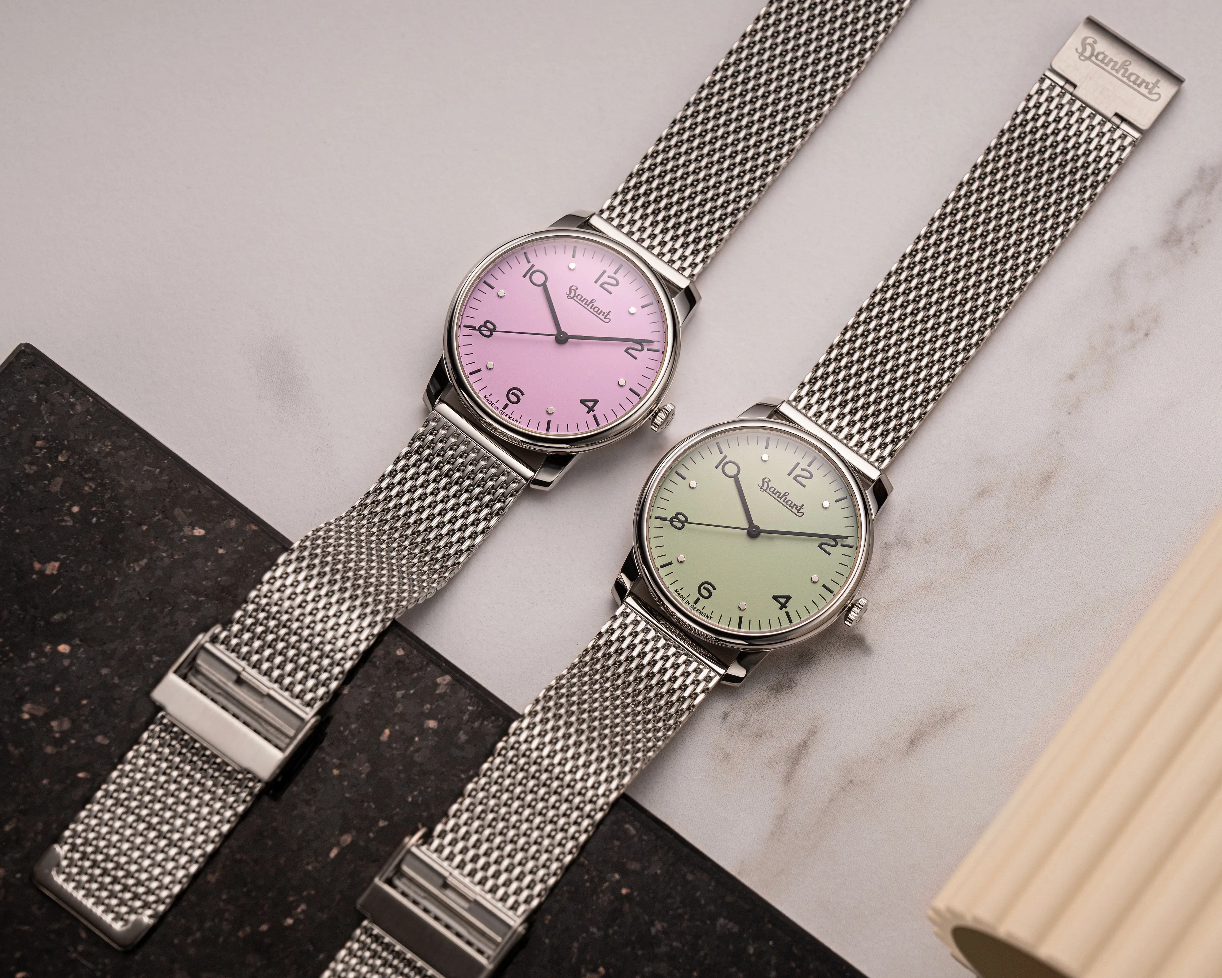 Two wristwatches with silver mesh bands, one with a pink dial and the other with a light green dial, on a marble surface.
