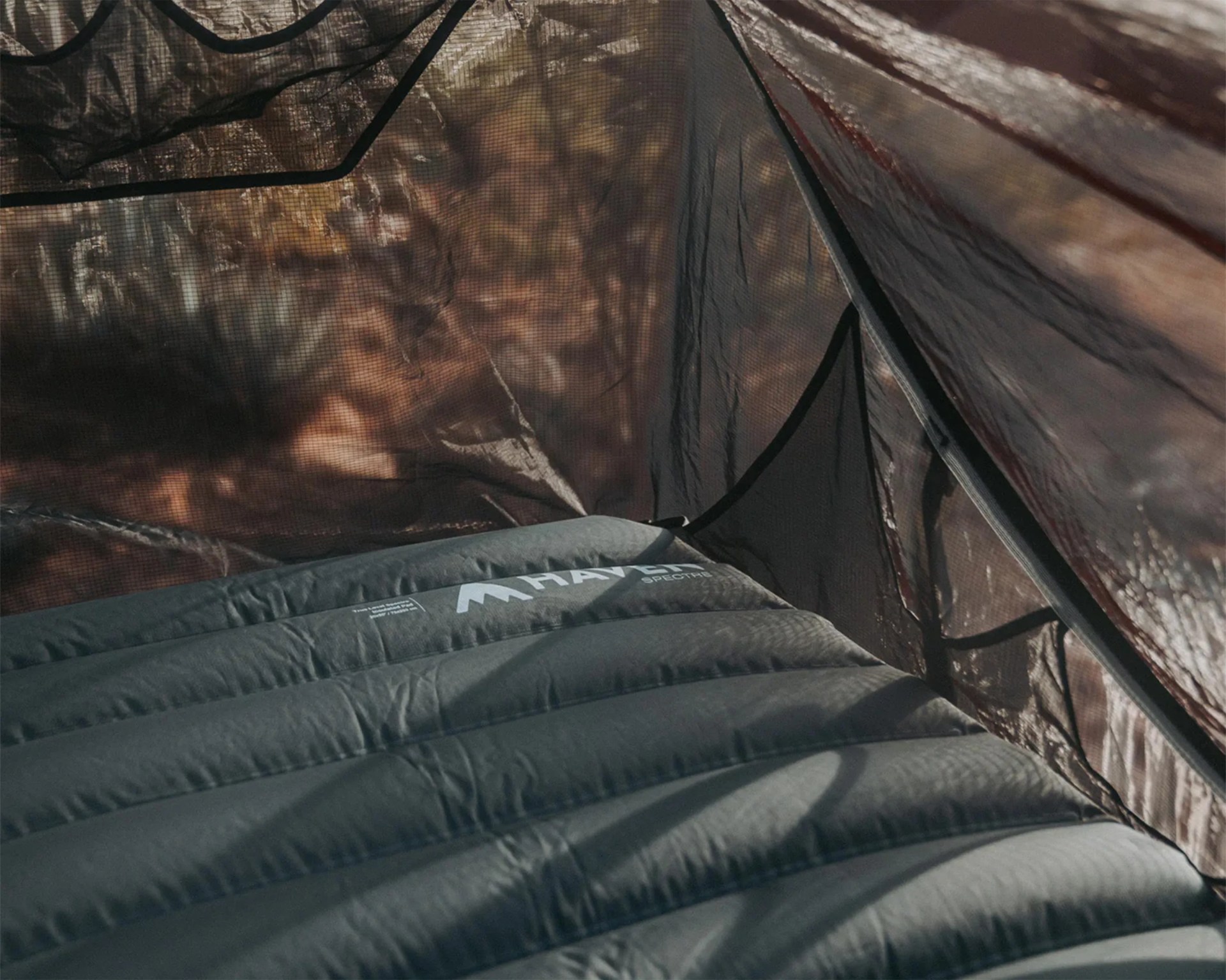 Dark gray inflatable sleeping pad inside a tent with mesh walls and autumn foliage visible outside.