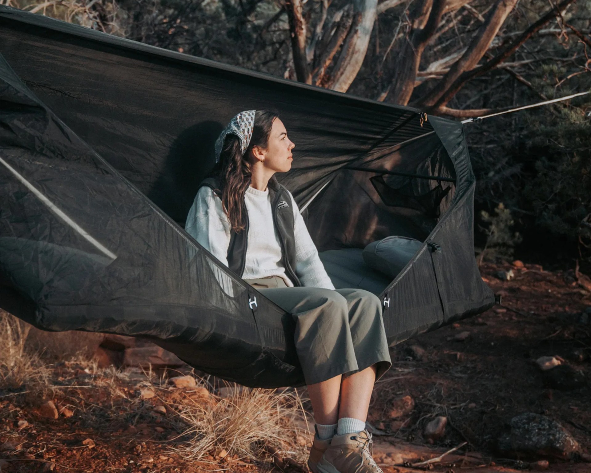 Person sitting in a black suspended hammock tent with mesh sides in a wooded outdoor setting.
