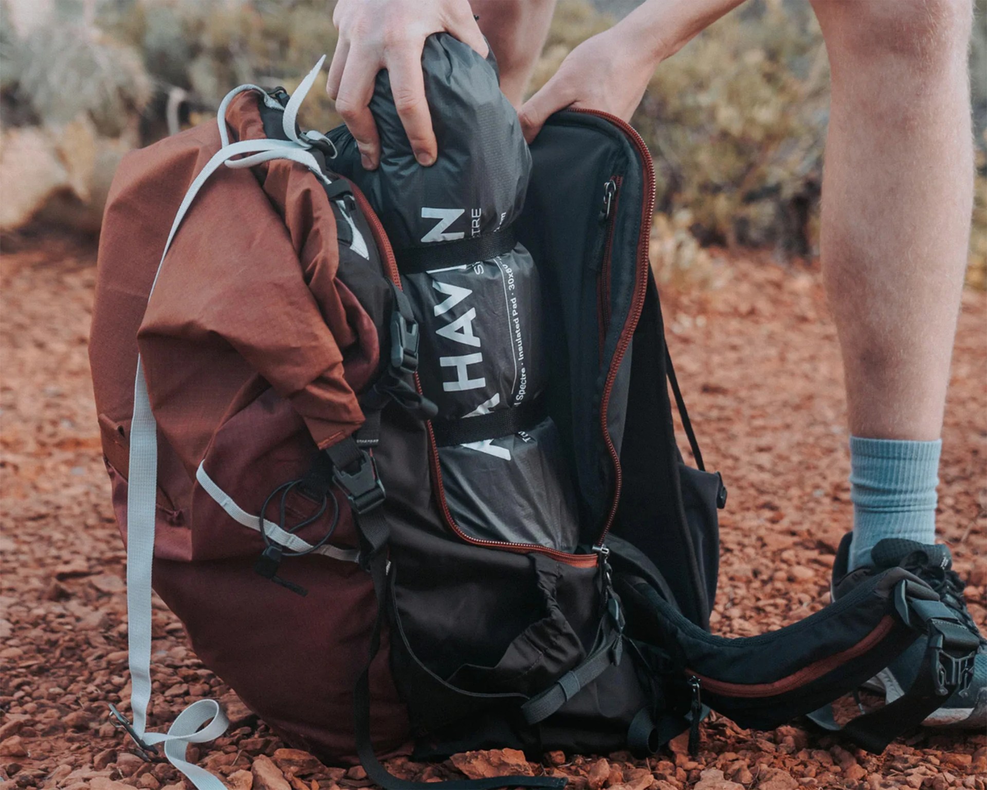Brown and black hiking backpack with gray straps being packed with a rolled black sleeping bag on rocky ground.