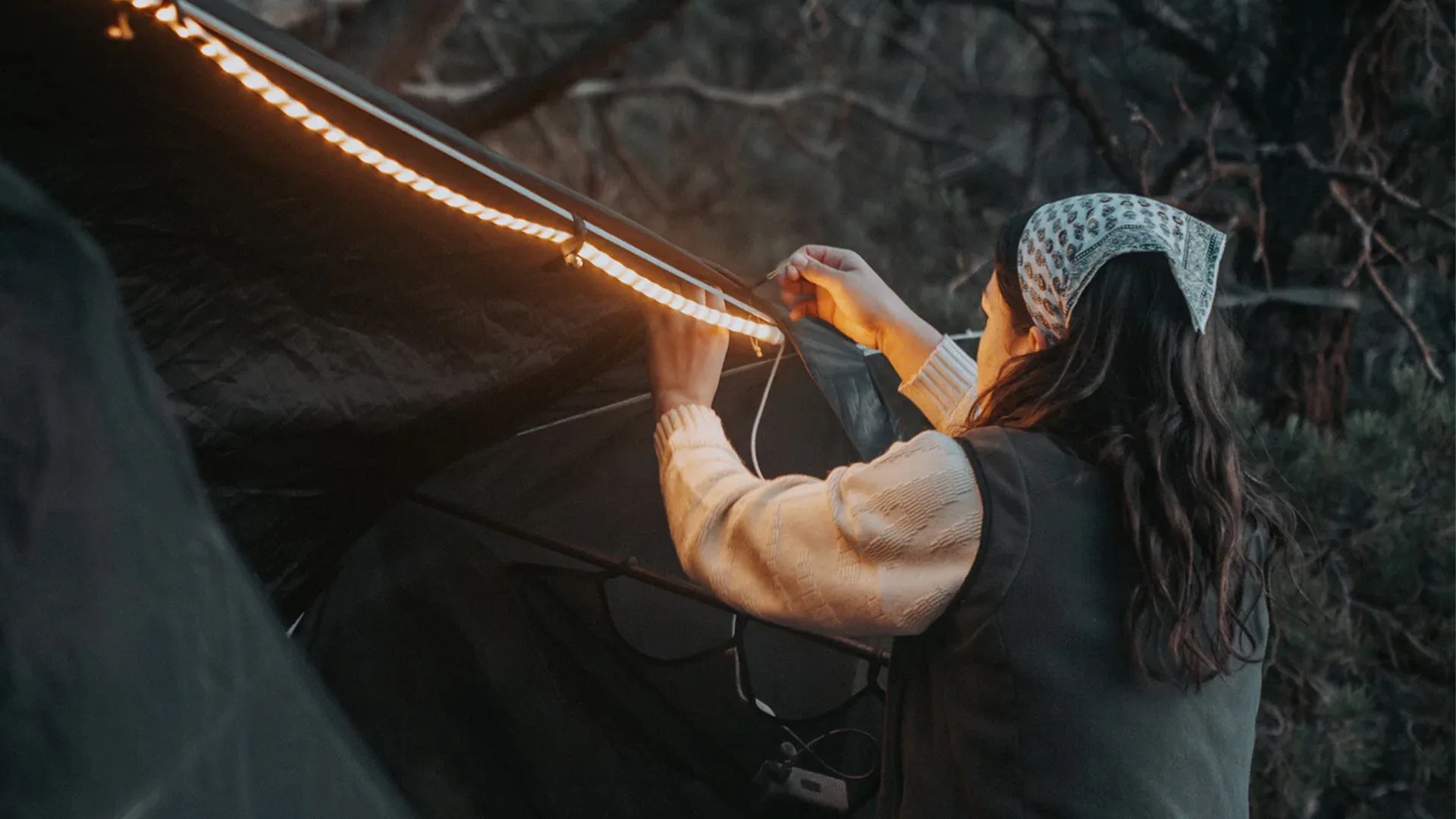 Person wearing a patterned bandana and sweater attaching a warm LED strip light to a tent at dusk.