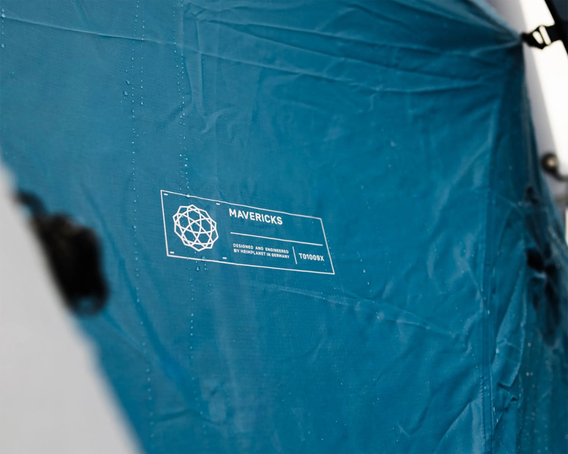Blue fabric with white "Mavericks" logo and text, showing water droplets on the surface.