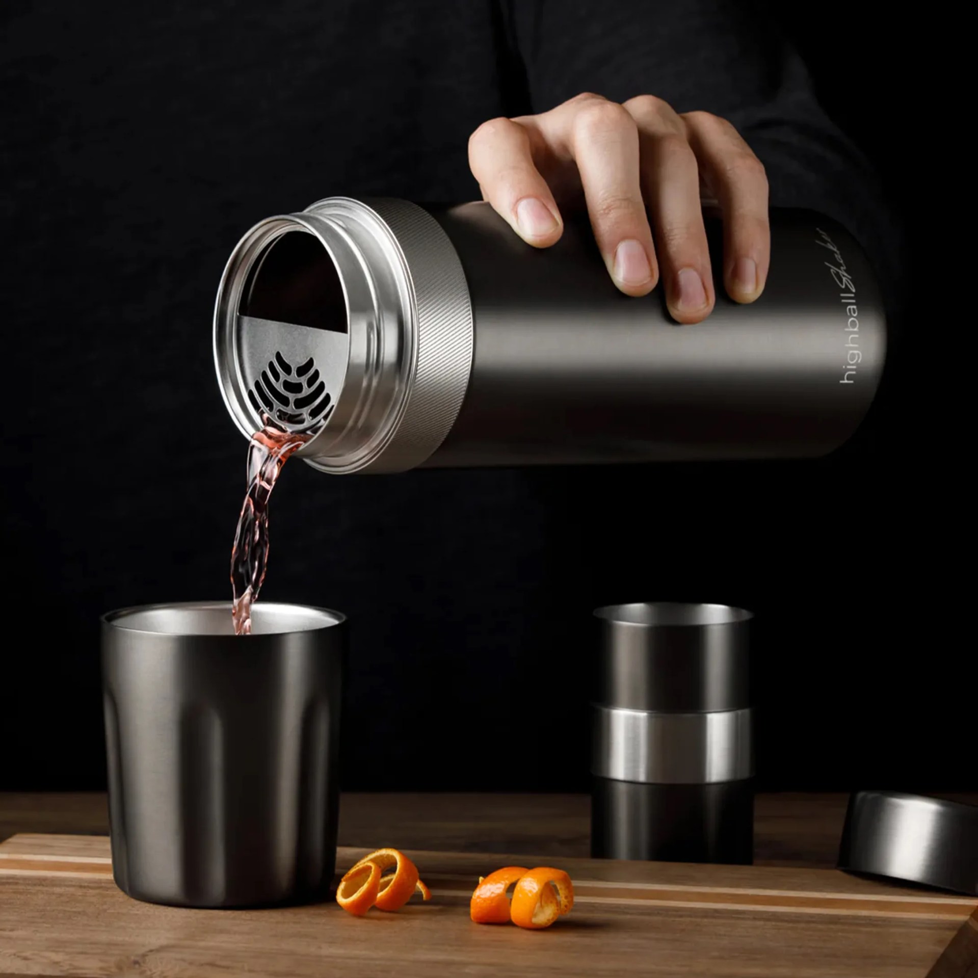 Matte black stainless steel cocktail shaker pouring liquid into a matching cup on a wooden surface with orange peels.