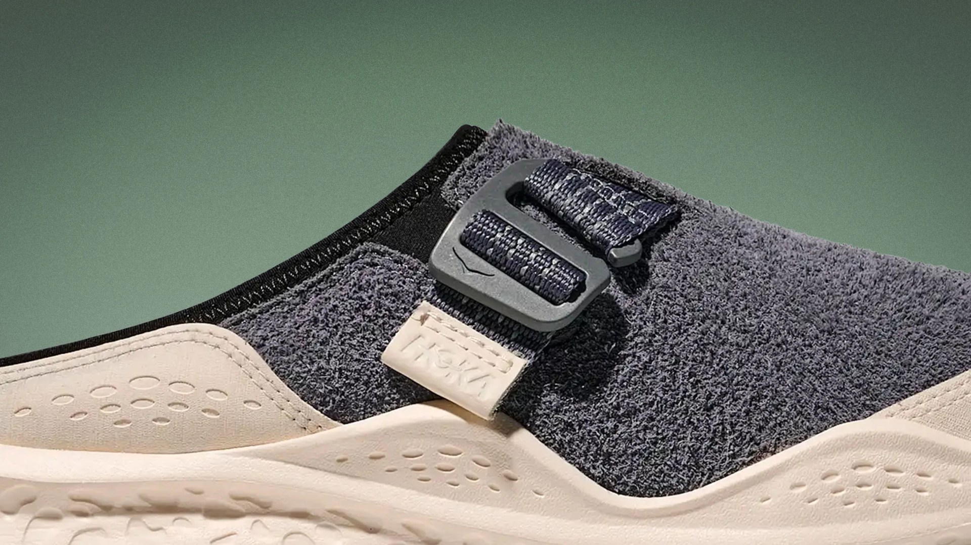 Close-up of a gray textured slip-on shoe with a beige sole and adjustable strap buckle.