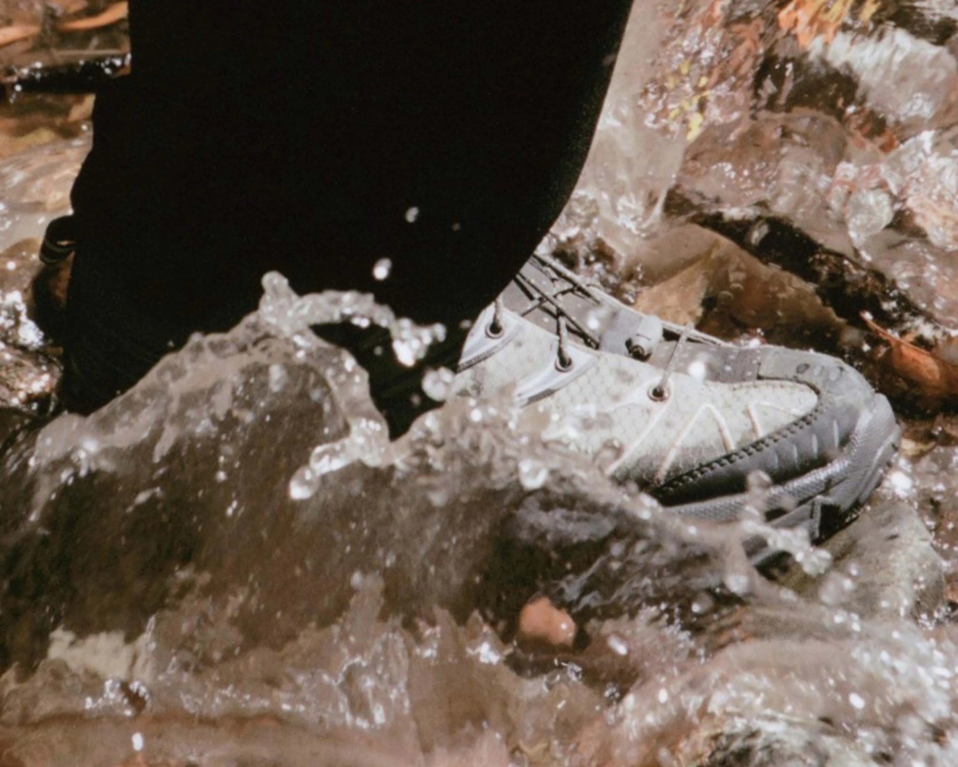 Gray and black hiking shoe splashing through shallow water over rocks.
