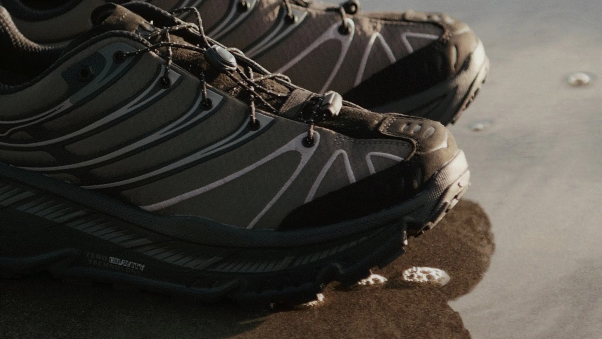 Pair of dark gray and black hiking shoes with reflective stripes on wet sandy ground.