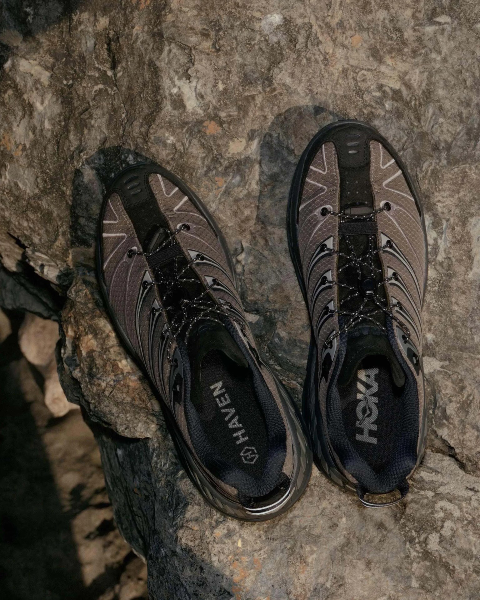 Pair of gray and black Hoka running shoes with Haven branding, placed on a rocky surface.