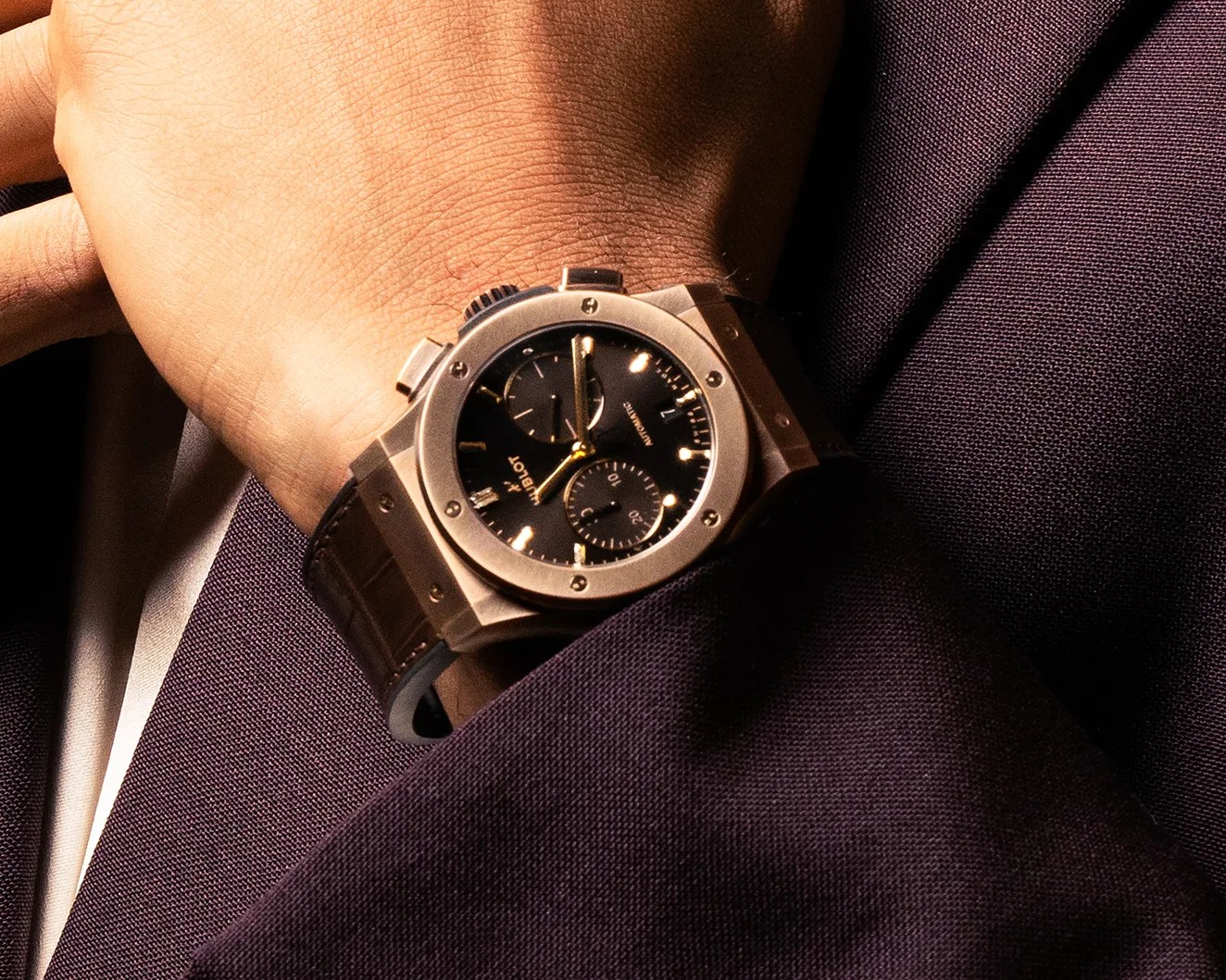 Gold-toned Hublot wristwatch with black dial and leather strap worn on a wrist over a dark purple suit sleeve.