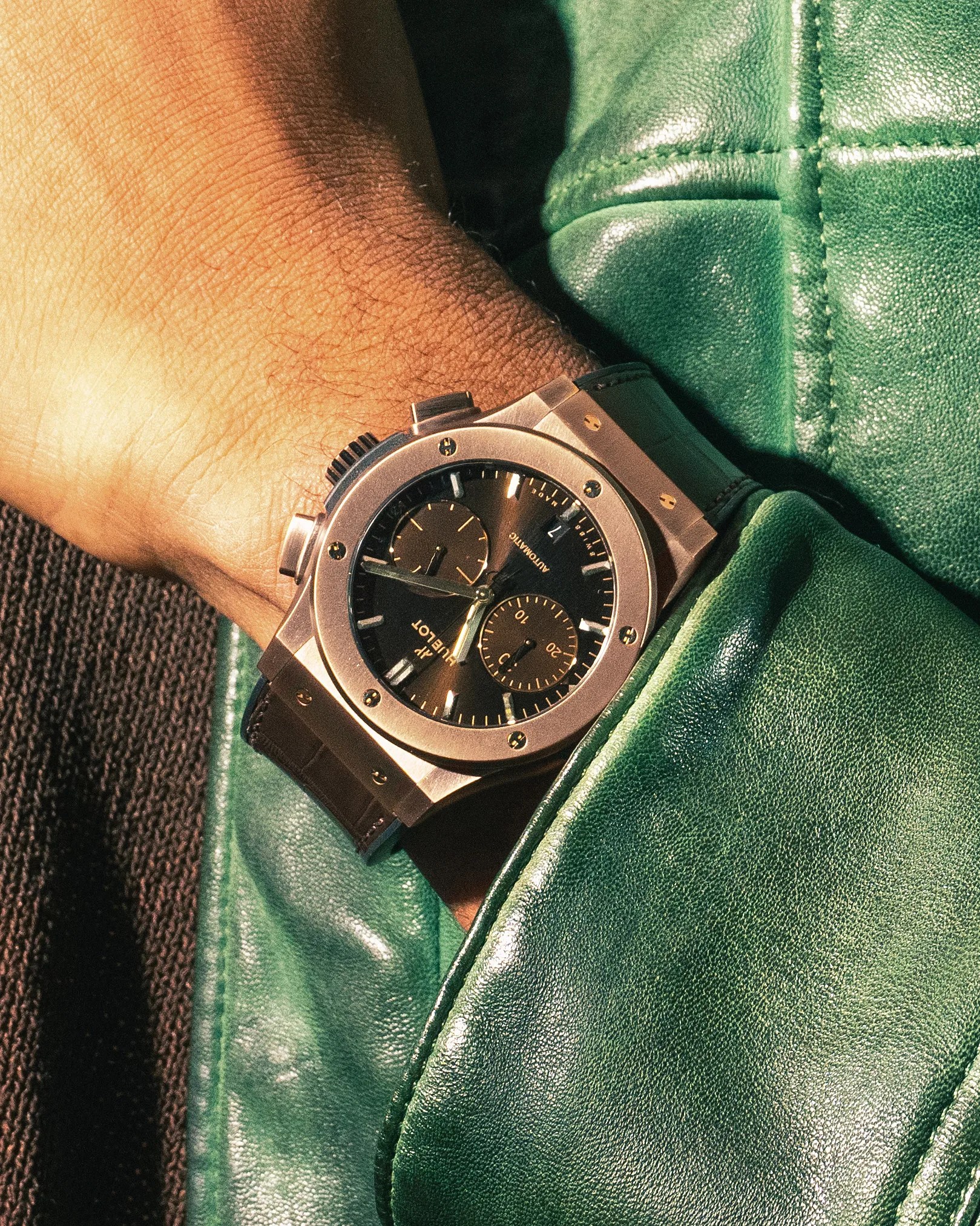Rose gold Hublot wristwatch with black dial and brown leather strap worn on a wrist over a green leather jacket.