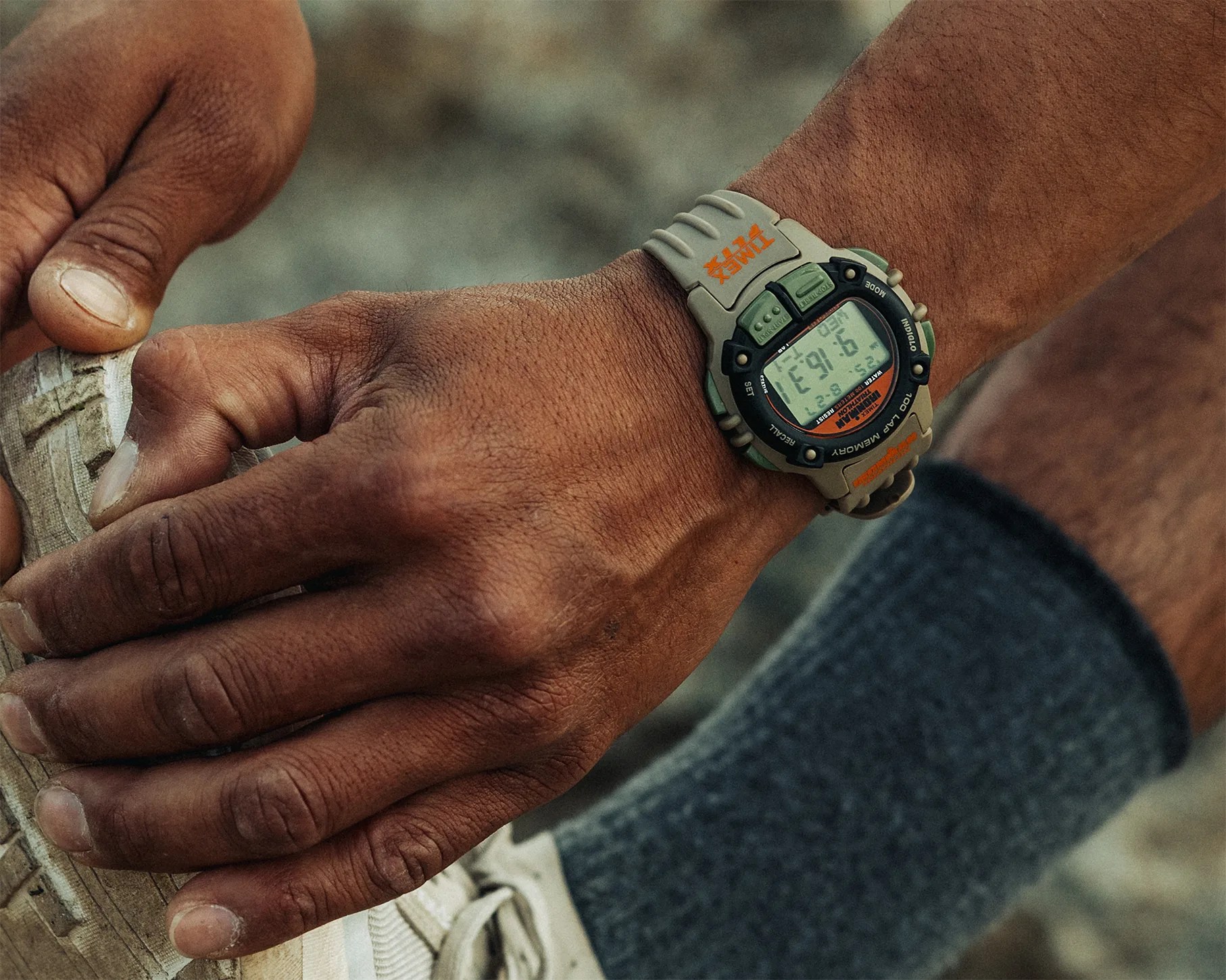 Close-up of a wrist wearing a tan and green Timex digital watch with an orange logo, holding a white sneaker.