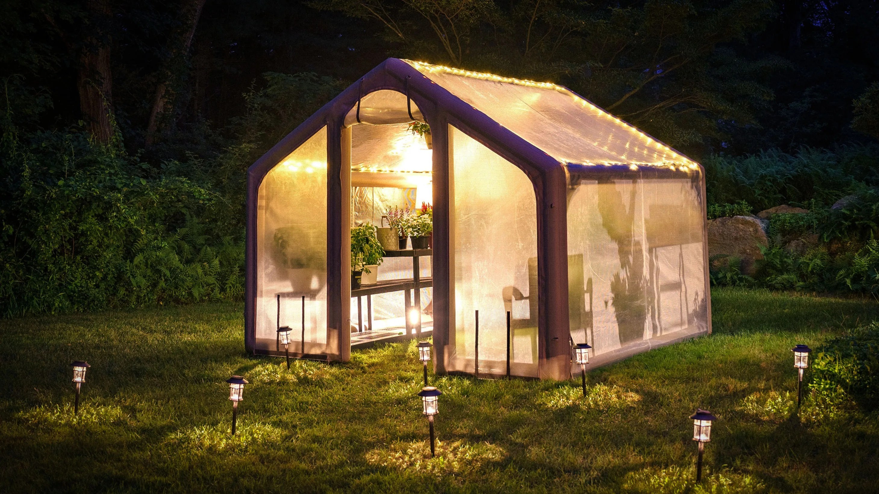 Illuminated small greenhouse with string lights and plants inside, surrounded by solar garden lights on grass at night.