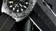 Close-up of a black dive watch with yellow accents, a textured bezel, and a black fabric strap with yellow stitching.