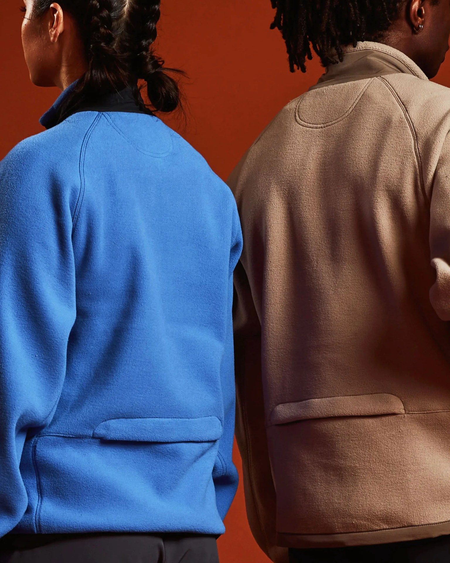 Two fleece jackets, one bright blue and one light brown, shown from the back on people against a rust-colored background.