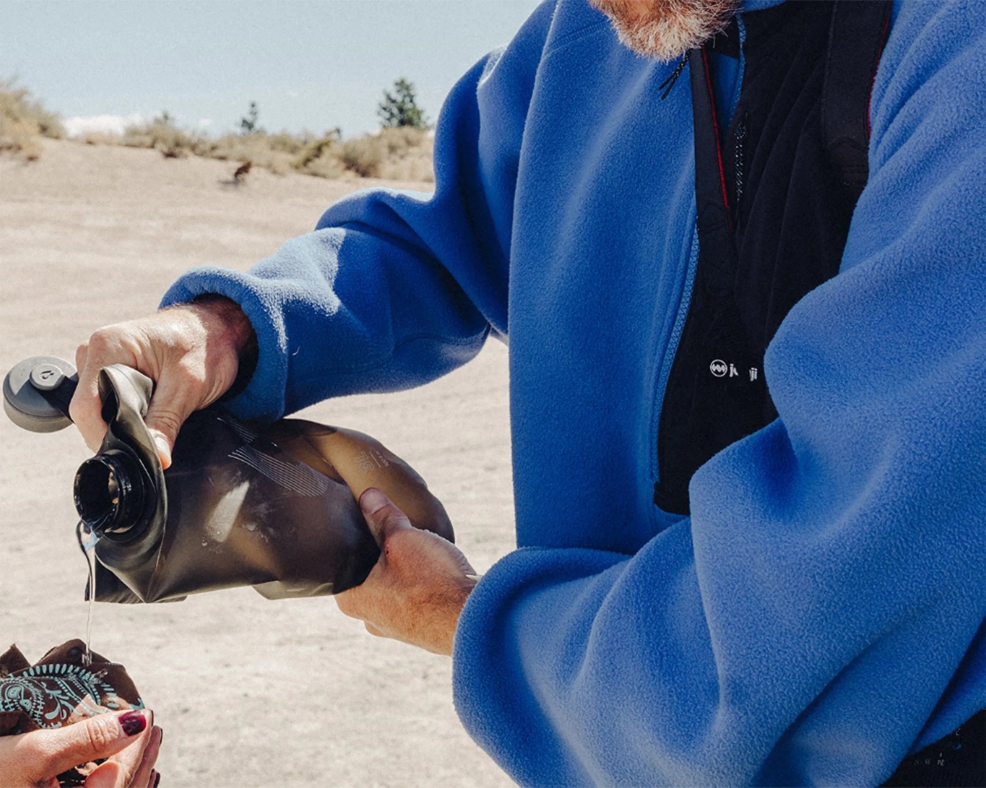 Person in a blue fleece jacket pouring water from a black collapsible water bottle outdoors.