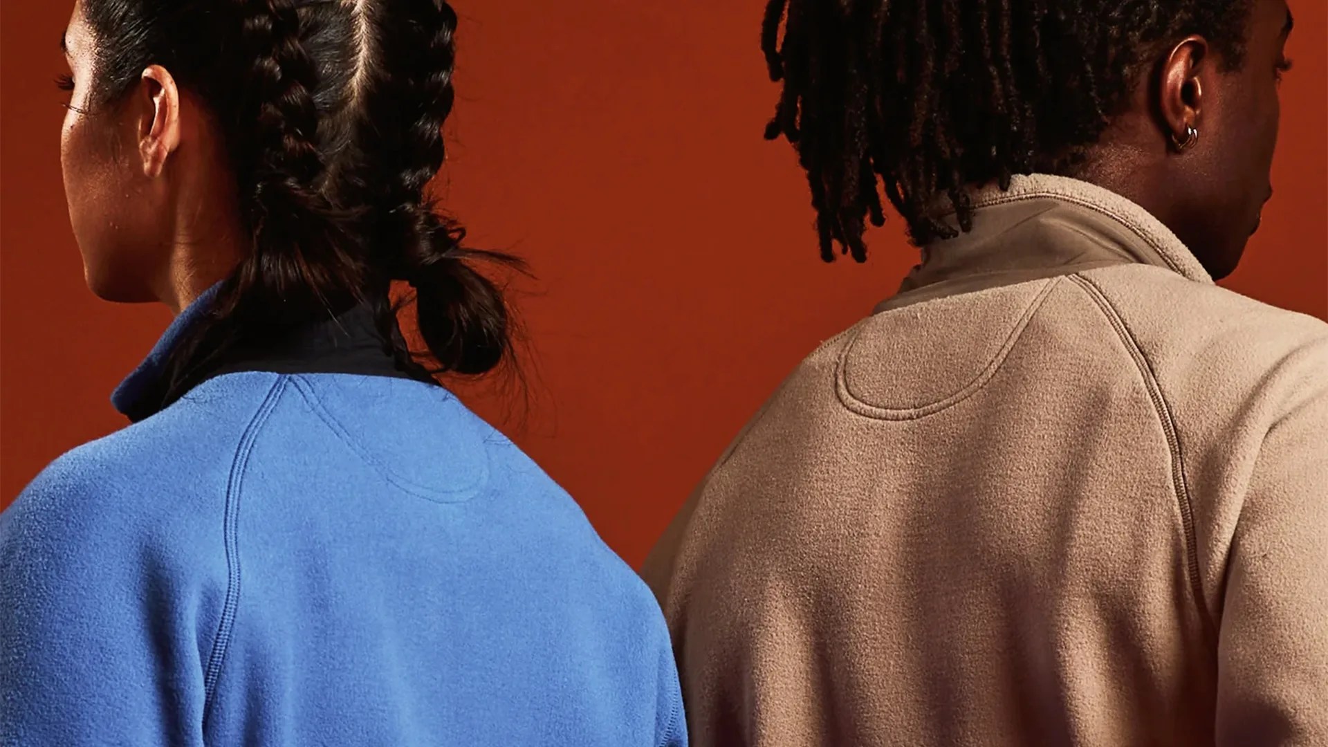 Two people wearing fleece jackets, one in light blue and the other in beige, shown from the back against a brown background.
