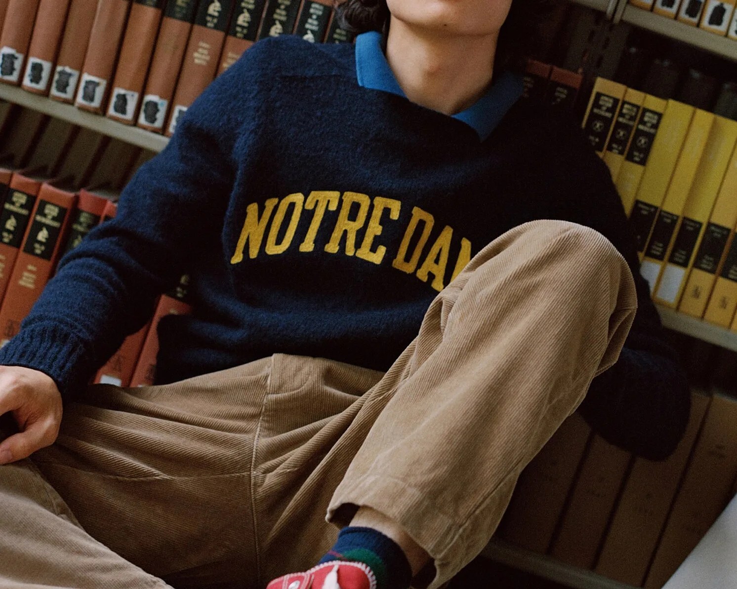 Person wearing a navy sweater with "NOTRE DAME" in yellow, tan corduroy pants, and red sneakers, sitting in front of bookshelves.