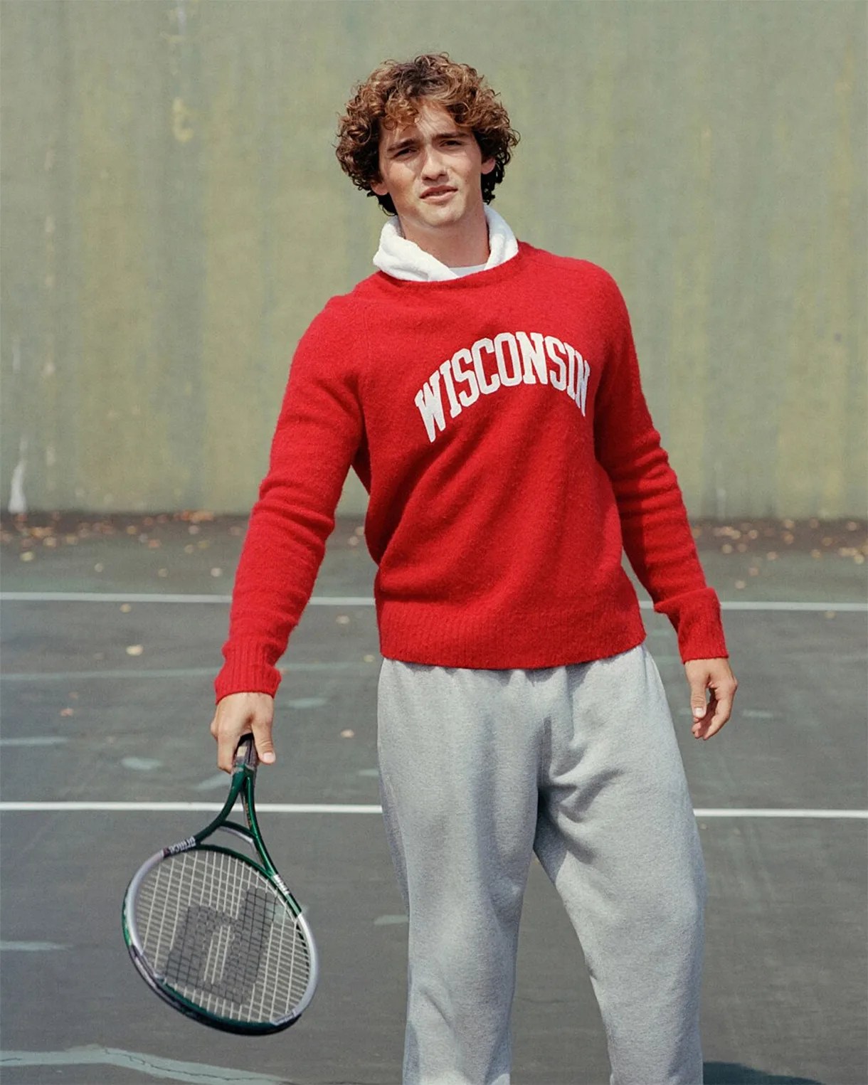 Person wearing a red "WISCONSIN" sweater and gray sweatpants holding a tennis racket on a court.