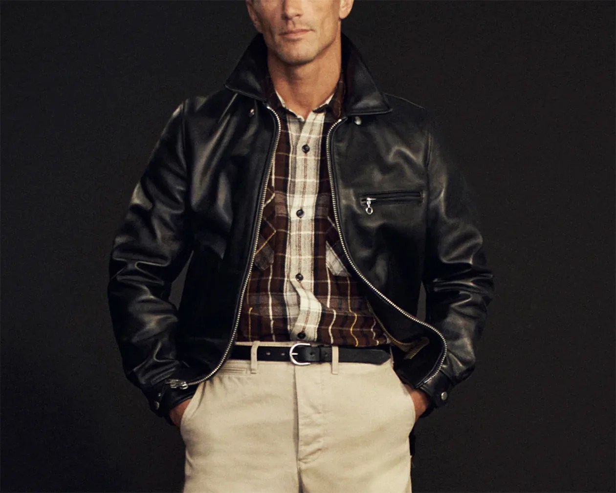Man wearing a black leather jacket over a brown and white plaid shirt with beige pants and a black belt.
