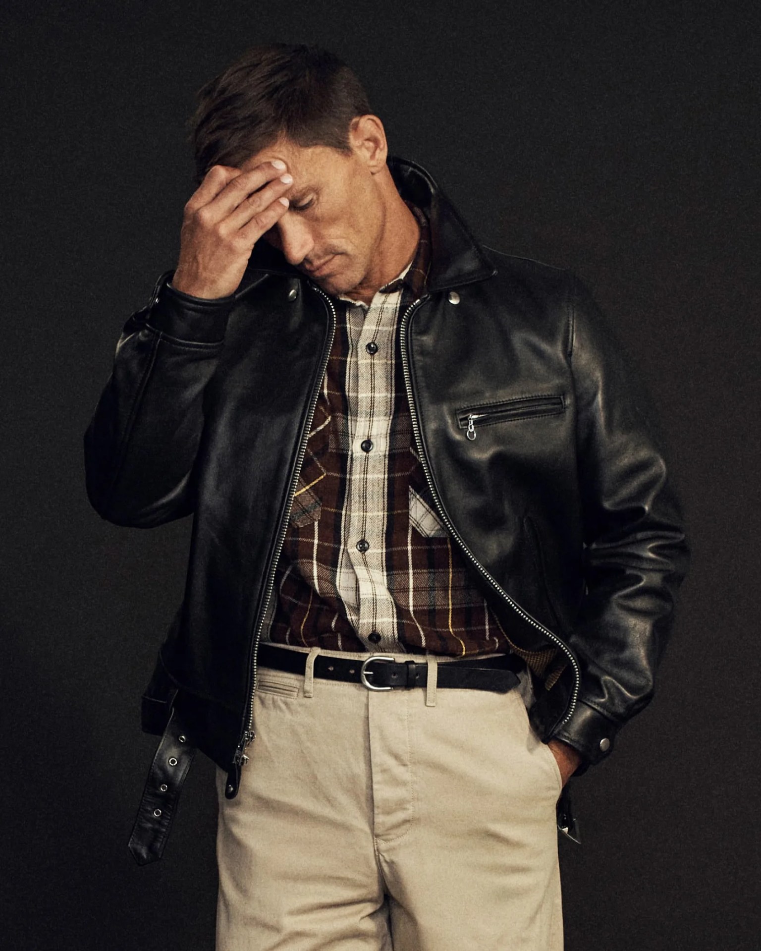 Man wearing a black leather jacket over a brown and white plaid shirt with beige pants and a black belt.