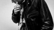 Close-up of a person wearing a black leather jacket over a plaid shirt, with their hand near their mouth.