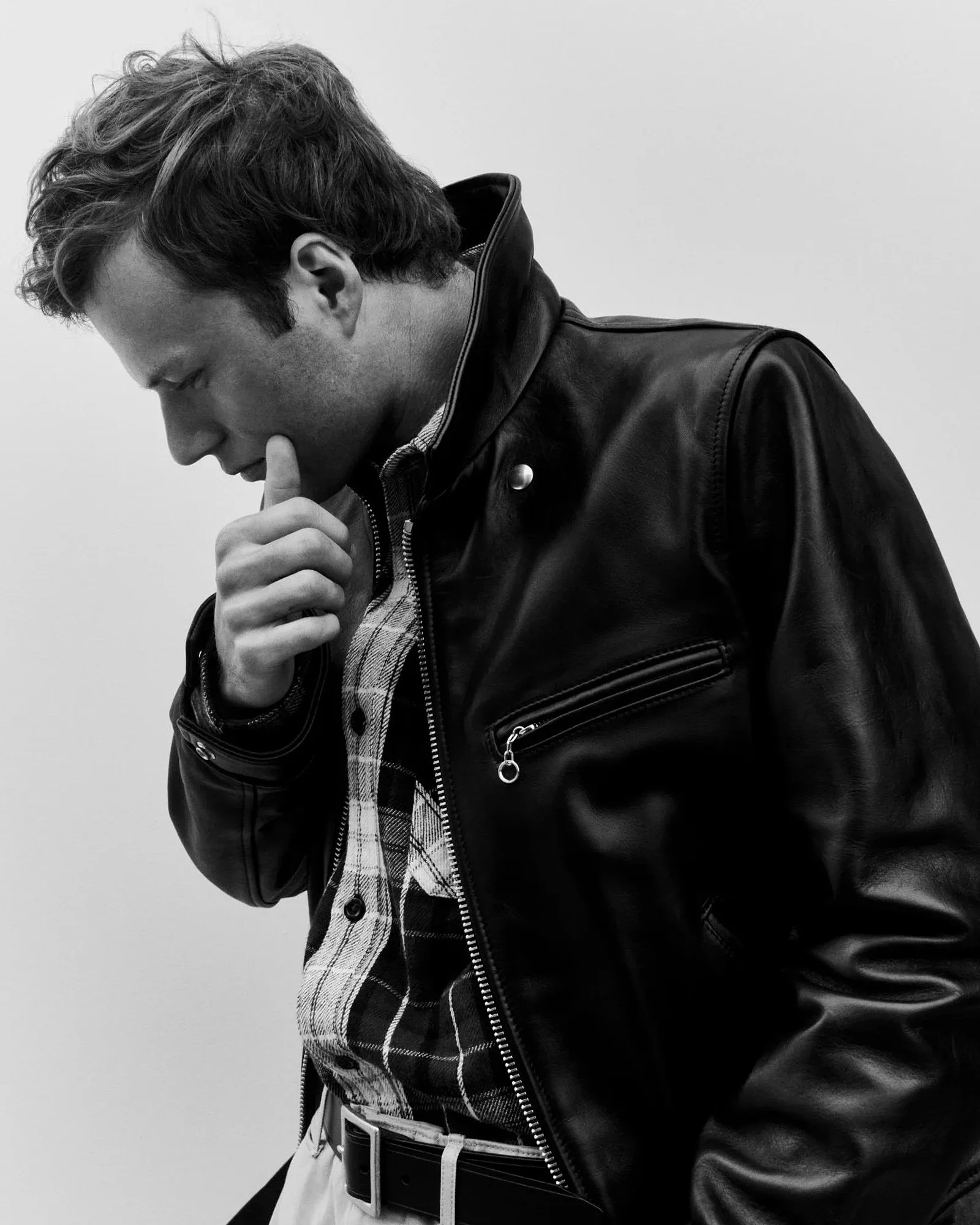 Man wearing a black leather jacket over a plaid shirt with a black belt, posing with hand near chin.