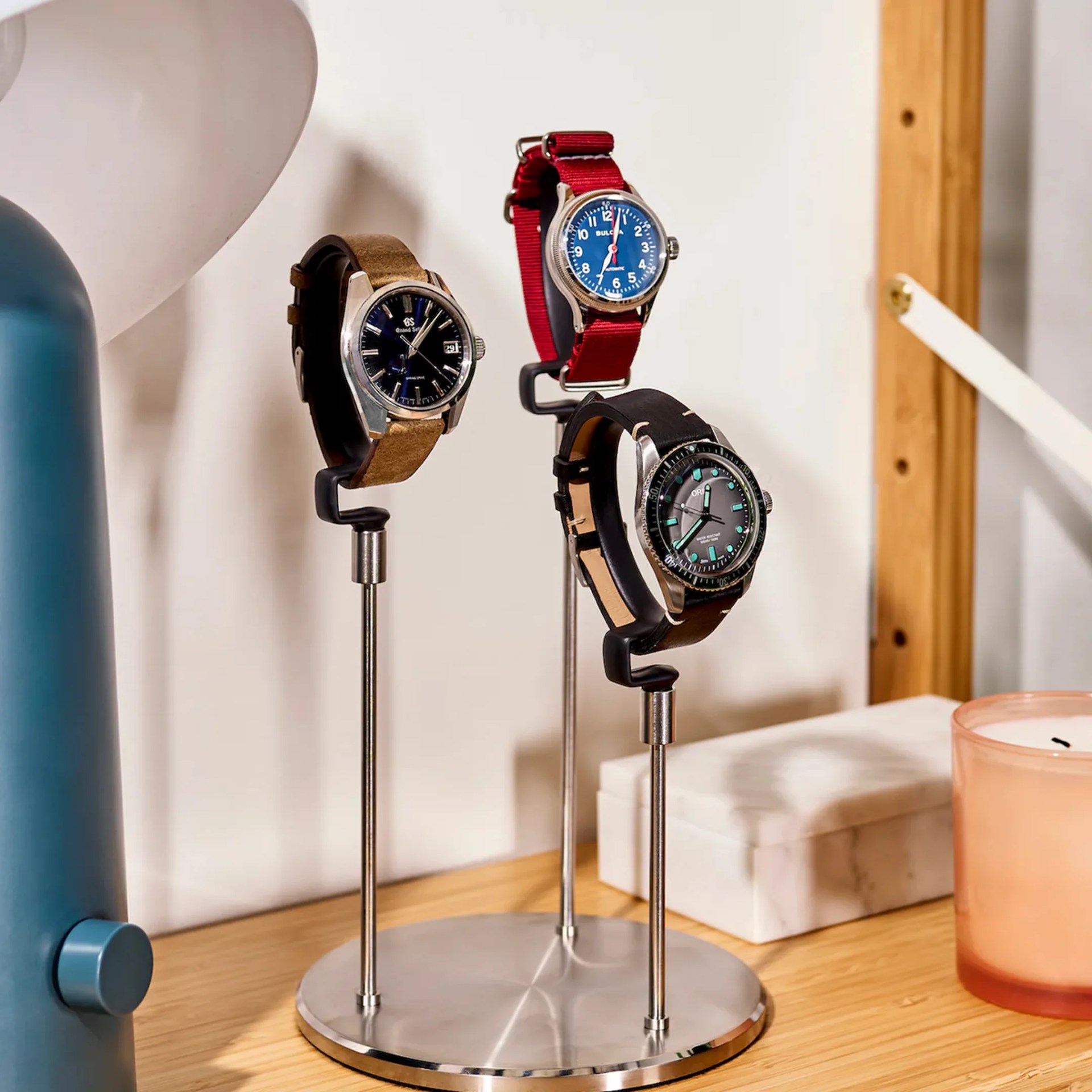 Three wristwatches with leather and fabric straps displayed on a three-prong metal stand on a wooden surface.