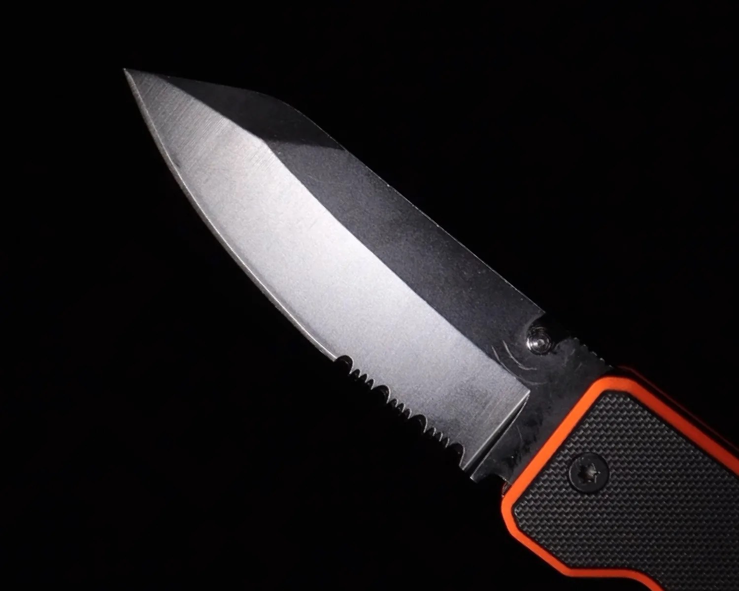 Partially serrated stainless steel knife blade with black and orange textured handle against a black background.