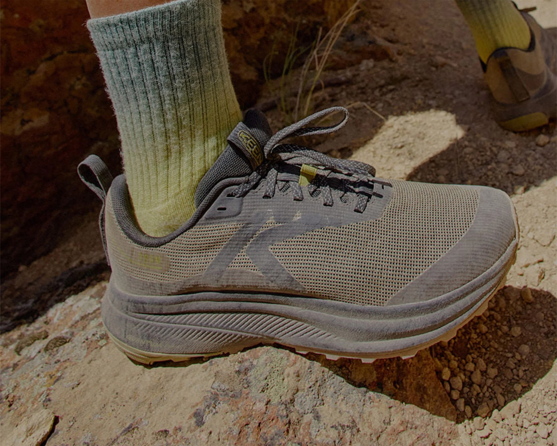 Gray mesh hiking shoe with black laces and thick sole worn with green socks on rocky terrain.