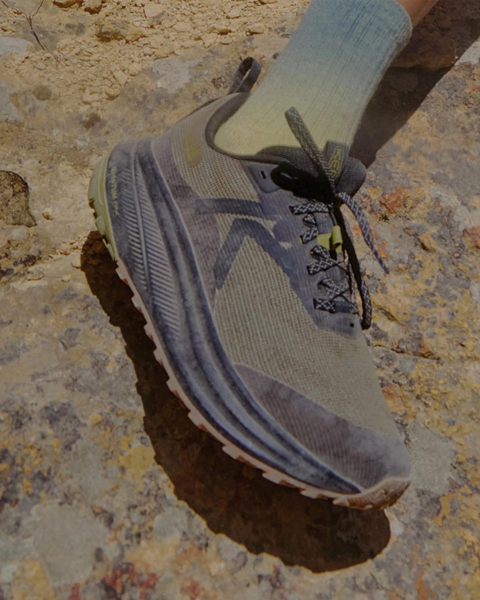 Gray and black athletic shoe with thick sole worn with light blue socks on rocky ground.