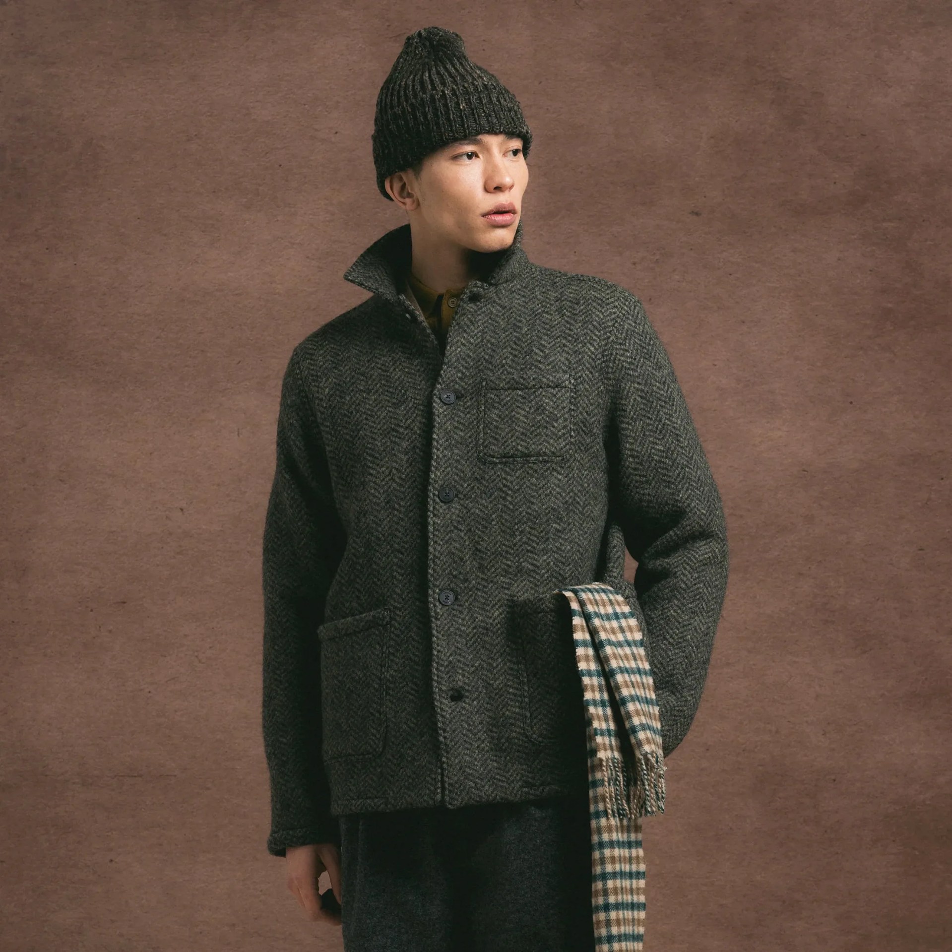 Man wearing a dark gray herringbone wool jacket and knit beanie holding a beige and green plaid scarf.