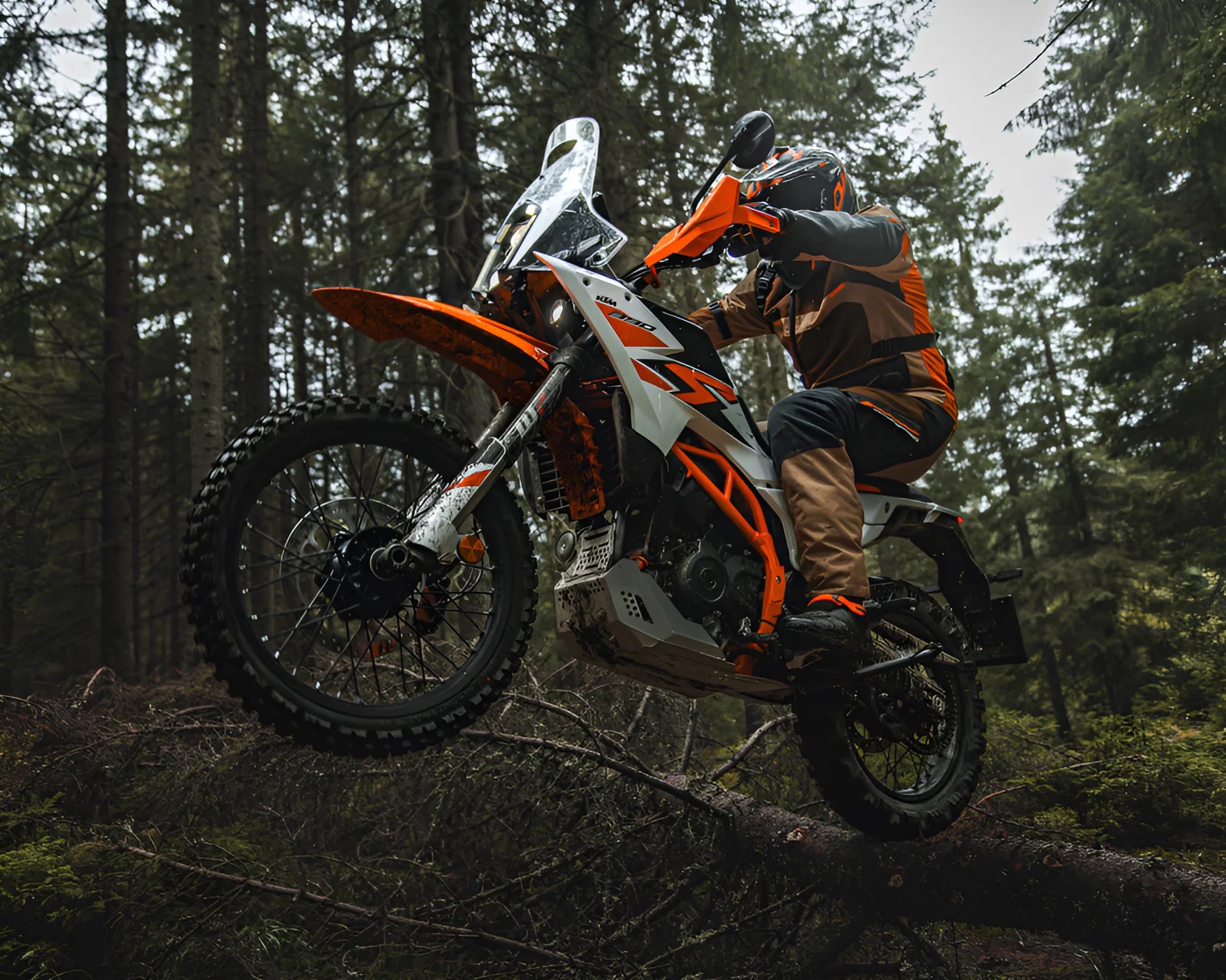 Orange and white KTM dirt bike with rider in matching gear jumping over a fallen tree in a forest.