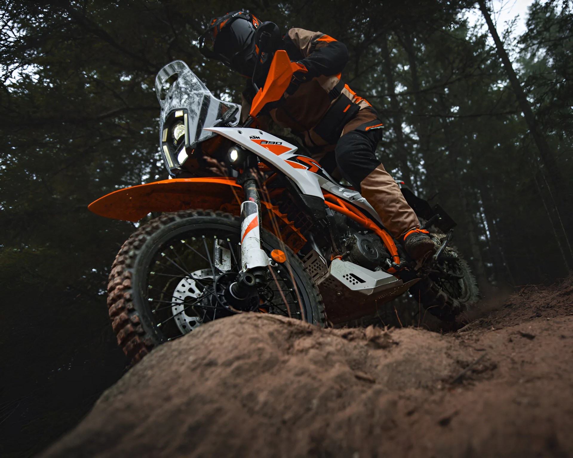Off-road motorcycle with orange and white body and knobby tires ridden on a dirt trail in a forest.