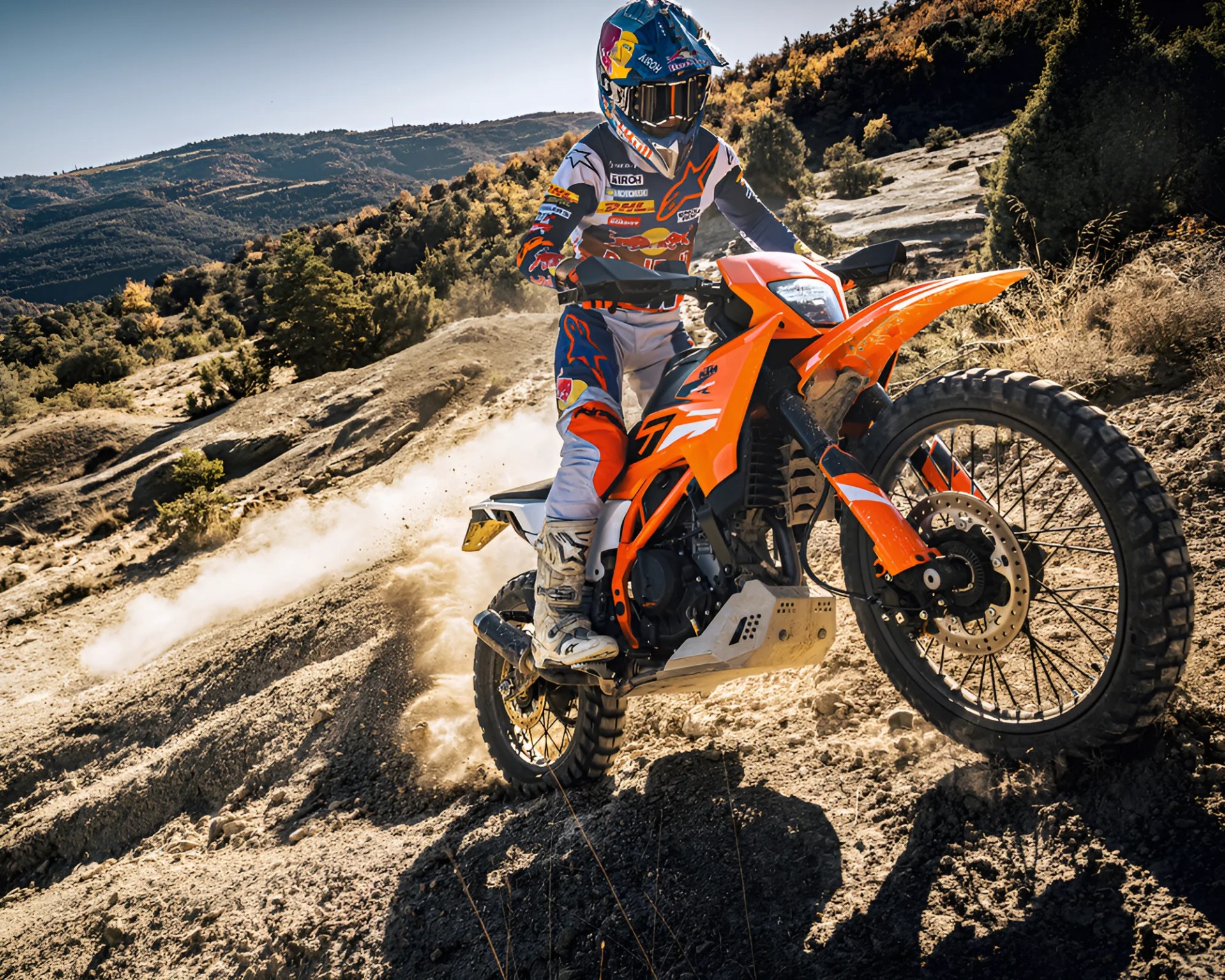 Rider in full gear on an orange KTM dirt bike kicking up dust on a rugged off-road trail.