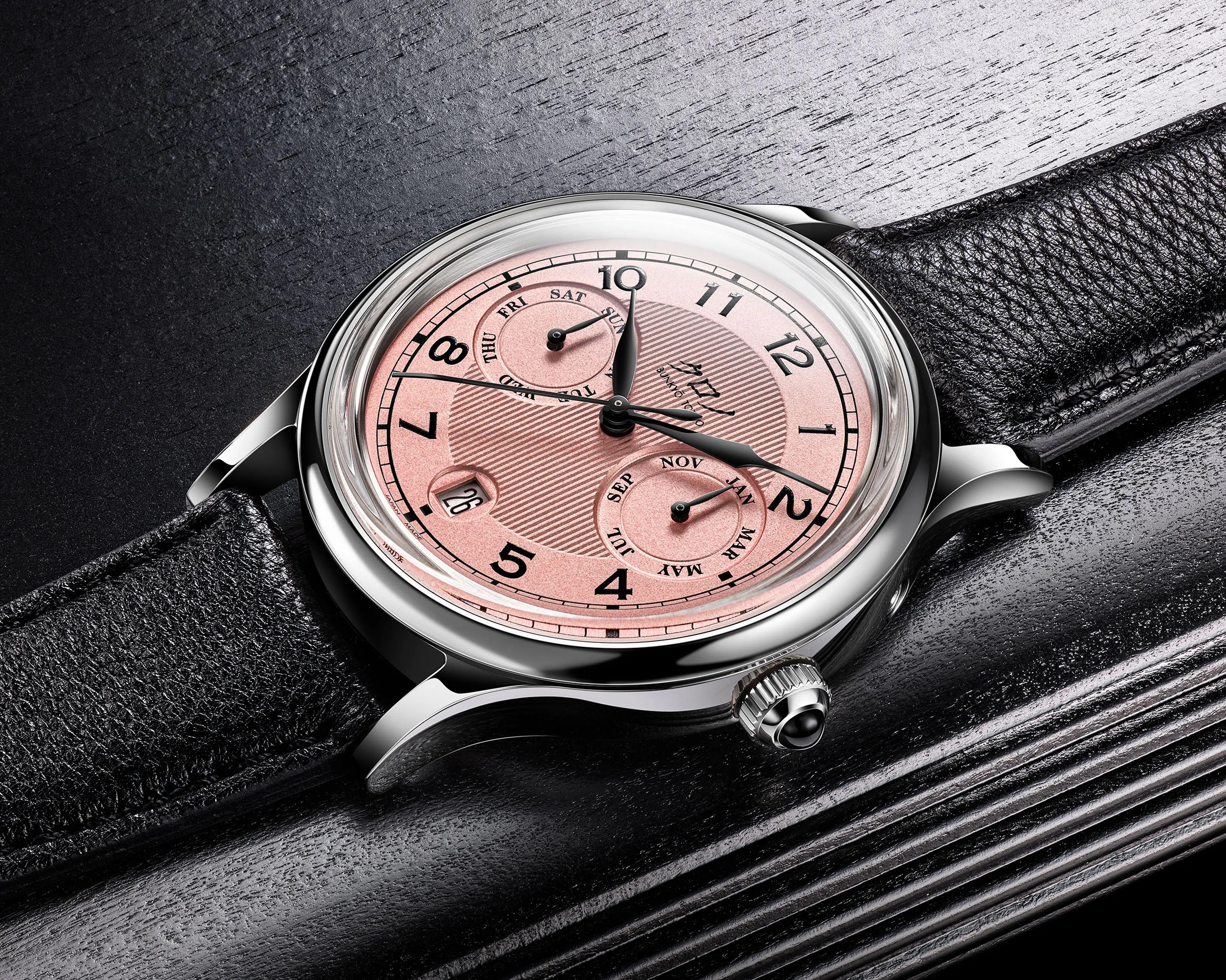 Silver wristwatch with a pink dial, black leather strap, and subdials for day and month on a black textured surface.