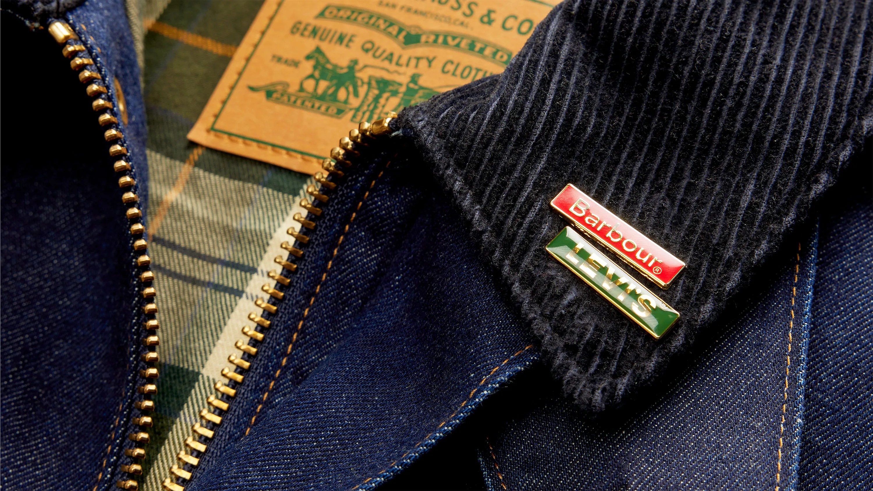 Close-up of a dark blue denim jacket with a brass zipper, black corduroy collar, and Barbour and Levi's pins attached.